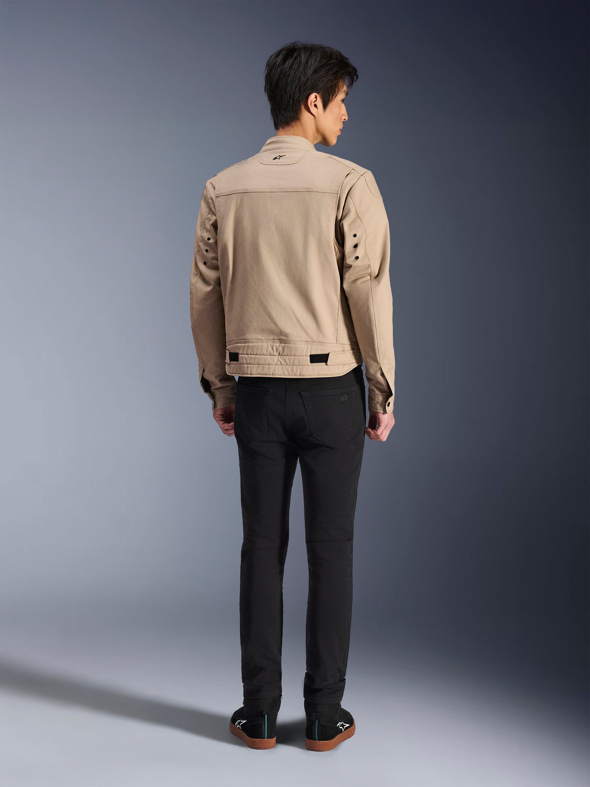 Rear view of a model wearing the Alpinestars Bruiser Jacket in brown, an urban motorcycle jacket with workwear-inspired design, featuring metal eyelet details on the back and waist adjusters, paired with black pants and sneakers, contemporary street riding style.