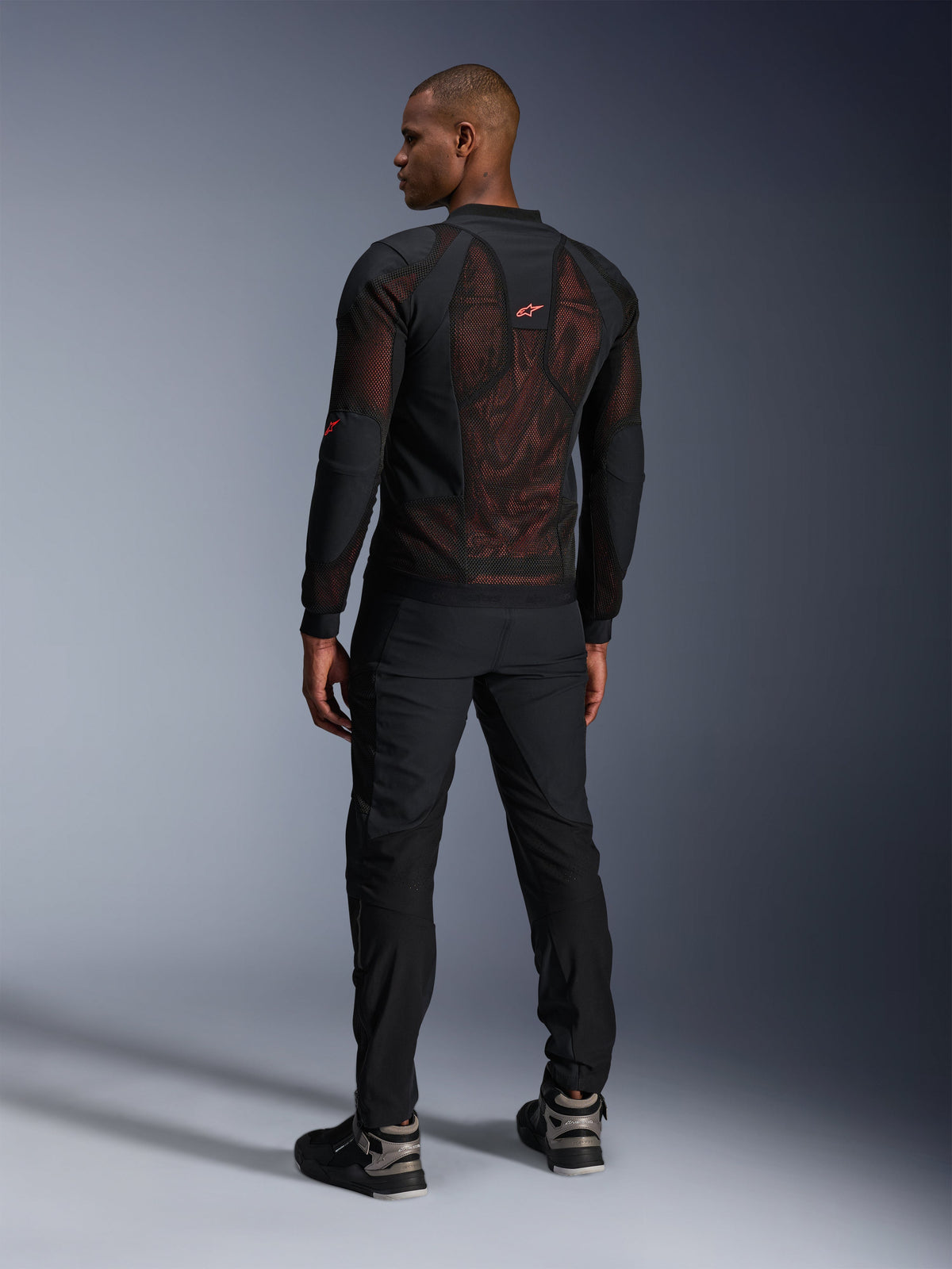 Alpinestars Troop-Air Pro Jacket in Black Red Fluo, rear view on a model, featuring a large 2D tech mesh back panel with visible red interior, paired with black technical riding pants and black/grey boots, modern urban armored motorcycle jacket design.