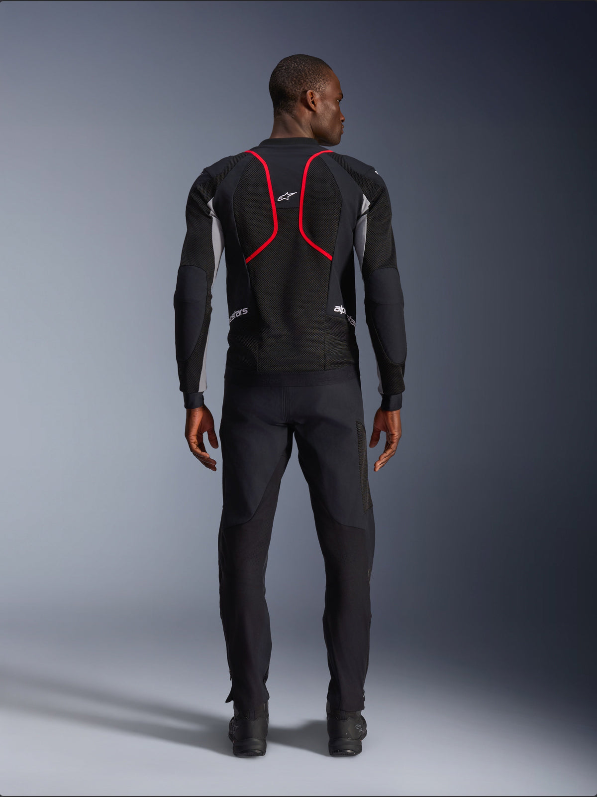 Model viewed from behind wearing the Alpinestars Troop-Air Pro Jacket, Black with Ash Gray and Bright Red accents, featuring technical mesh construction with prominent red piping on the back, Alpinestars logo on the upper back, paired with black motorcycle pants, premium armored air jacket for road riding