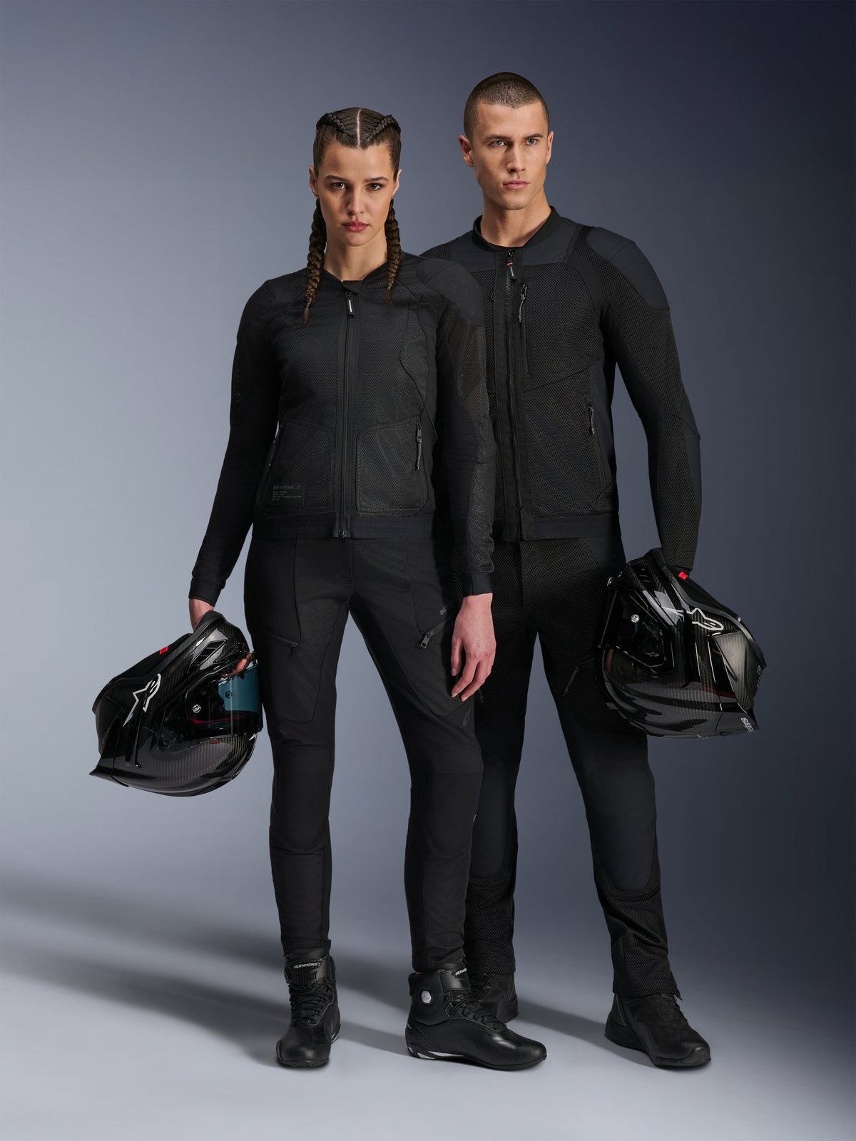 Modelos masculino y femenino con chaquetas Alpinestars Troop-Air Pro en negro, equipo de malla técnica blindada combinado con pantalones y botas negros a juego, cada uno sosteniendo un casco negro, mostrando ropa de motociclismo urbana coordinada.