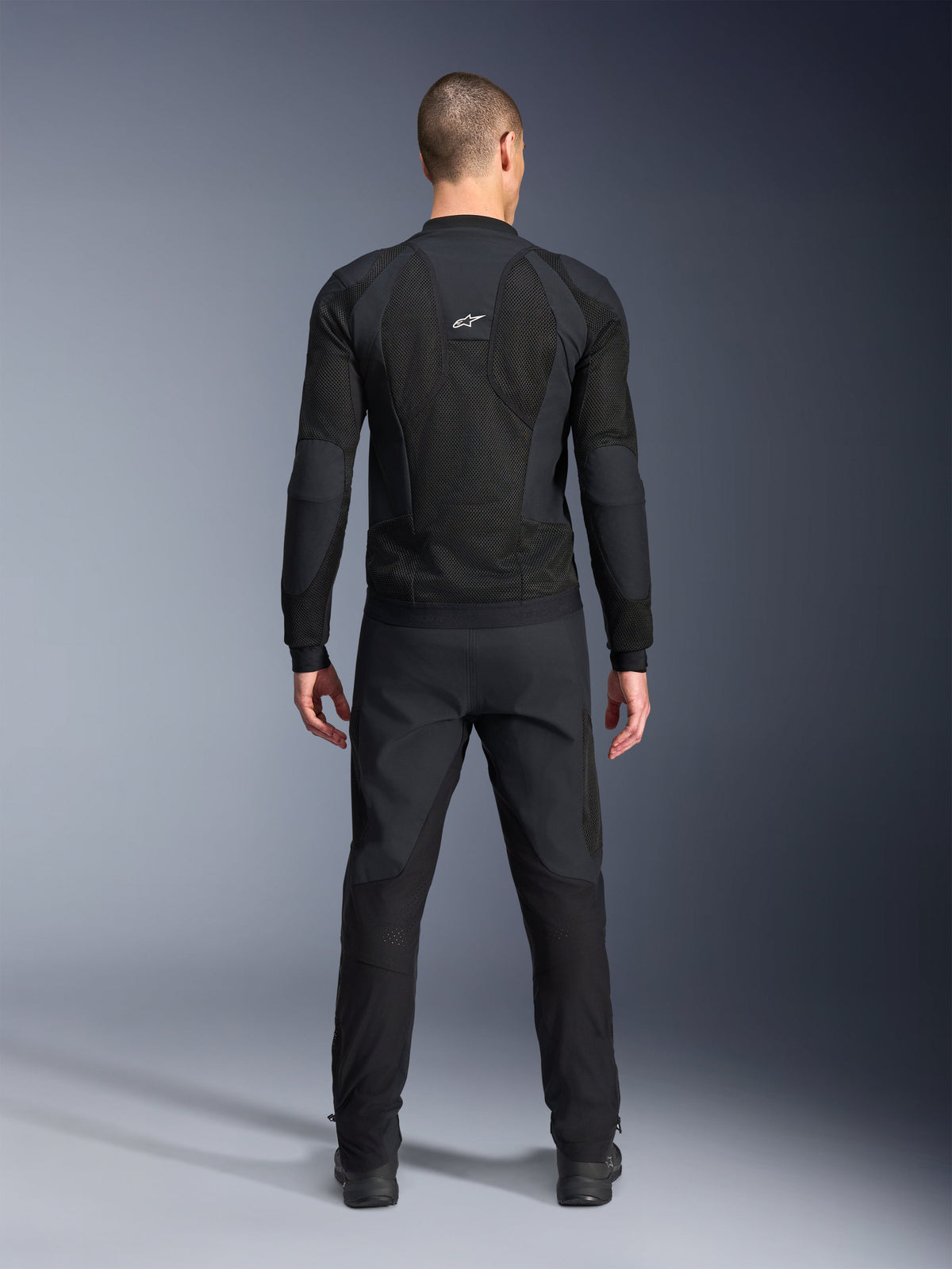 Chaqueta Alpinestars Troop-Air Pro, vista trasera, negra, chaqueta técnica de aire para motociclismo con grandes paneles de ventilación de malla y logotipo blanco de Alpinestars, combinada con pantalones negros, equipamiento de conducción urbana contemporánea
