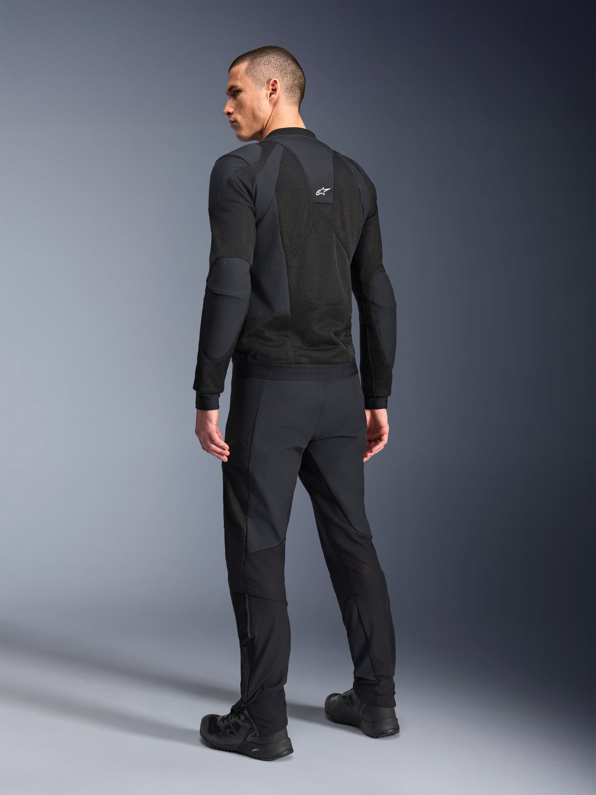 Chaqueta Alpinestars Troop-Air Pro, negra, chaqueta de aire técnica blindada vista desde la espalda, con amplios paneles de malla para el flujo de aire, combinada con pantalones técnicos negros y calzado a juego.