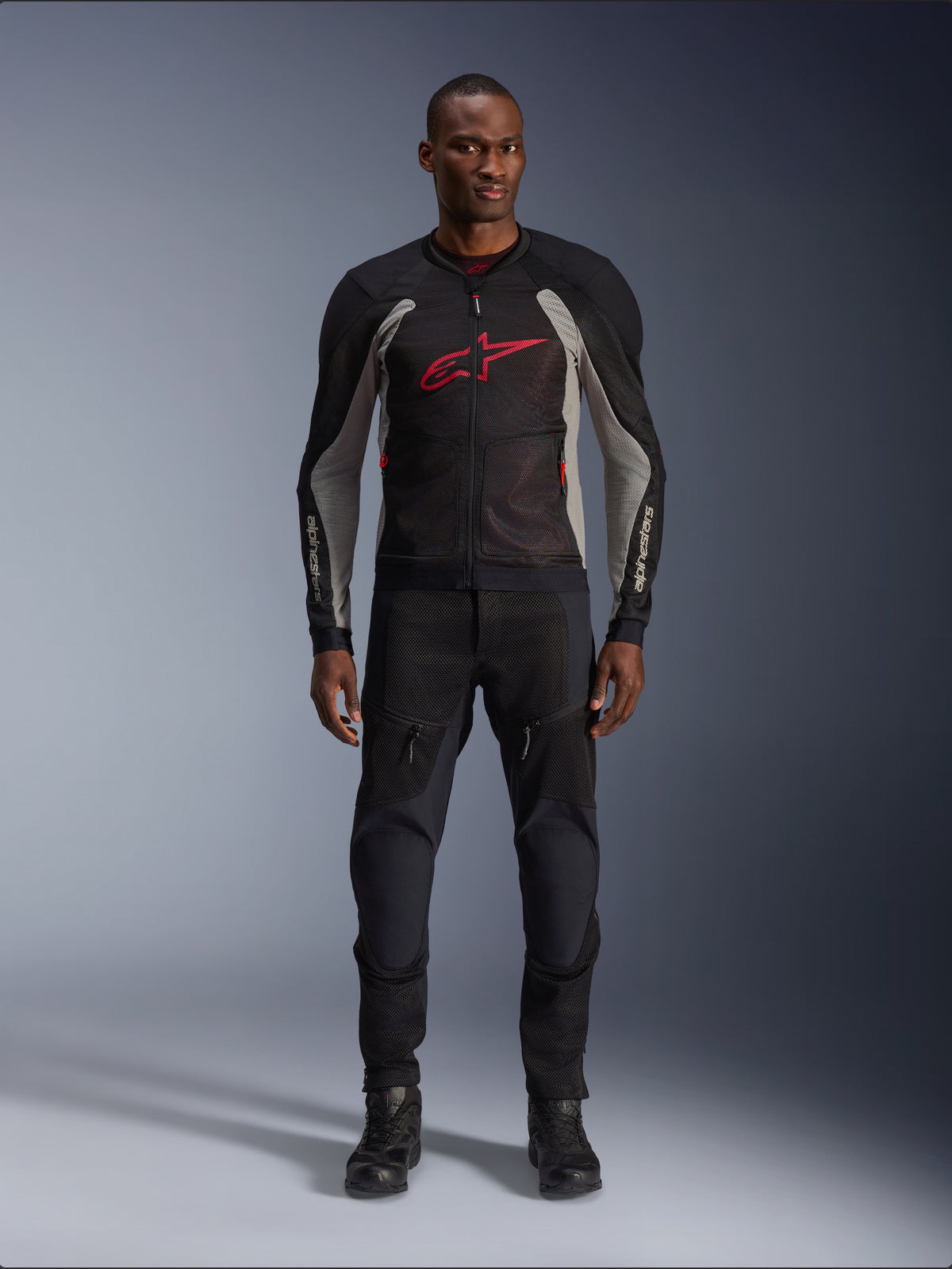 Modelo masculino con la chaqueta Alpinestars Troop-Air en color Negro Gris Ceniza y Rojo Brillante, chaqueta de malla técnica para moto con un prominente logotipo rojo en el pecho, combinada con pantalones y botas de moto negros, ropa de conducción urbana