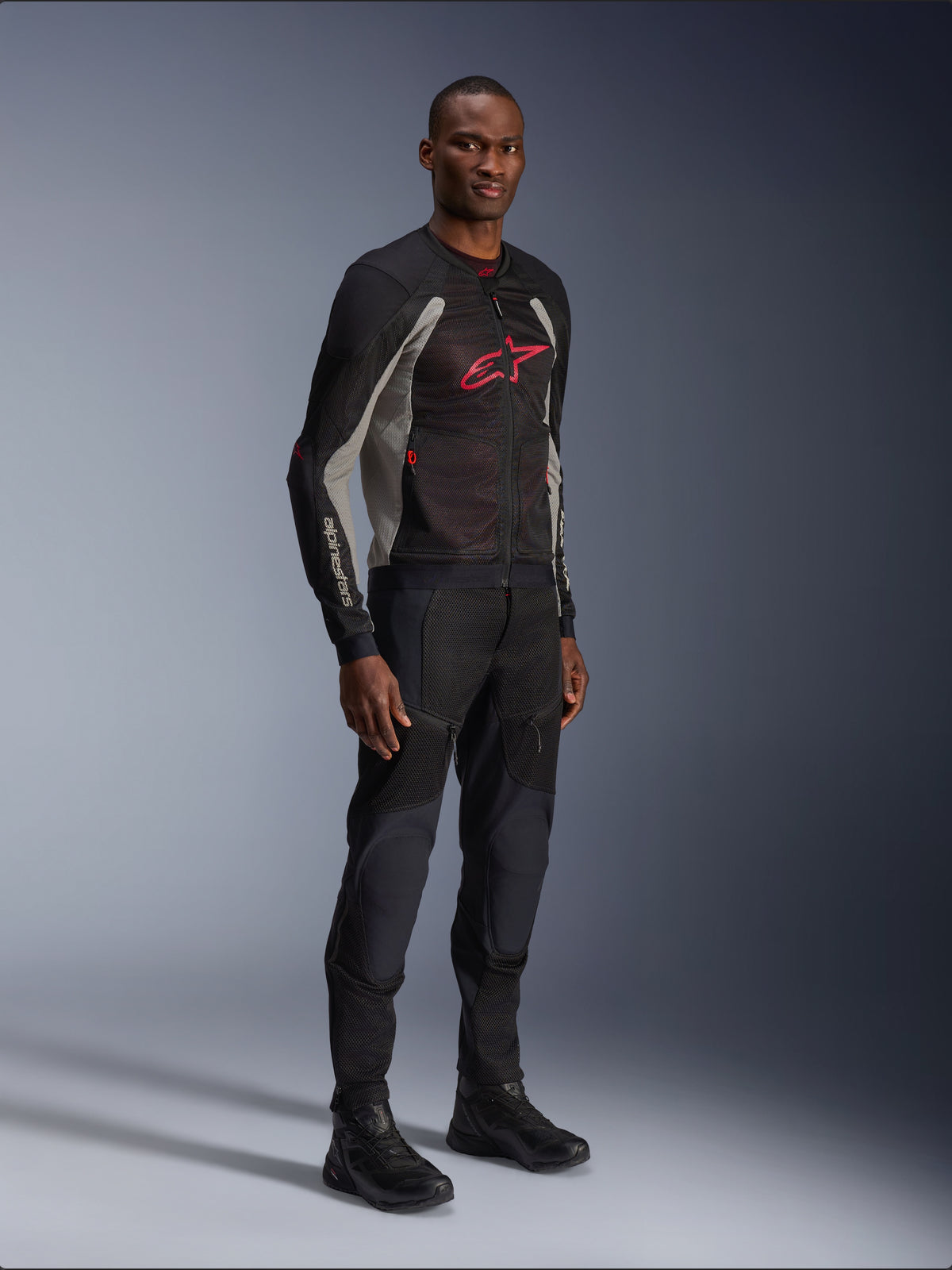 Chaqueta Alpinestars Troop-Air, chaqueta de malla técnica para motocicleta en negro, gris ceniza y rojo brillante, usada por un modelo con pantalones y botas negros a juego, con un logotipo de Alpinestars rojo en el pecho, equipamiento de conducción urbano contemporáneo.