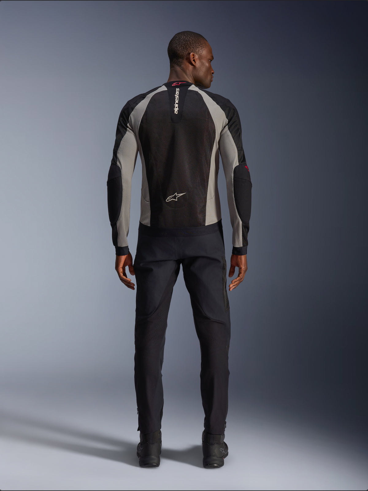 Chaqueta Alpinestars Troop-Air, chaqueta de moto negra, gris ceniza y rojo brillante, vista trasera en modelo masculino, construcción de malla ligera para ventilación, marca Alpinestars en el cuello y la parte inferior de la espalda, combinada con pantalones de conducción negros