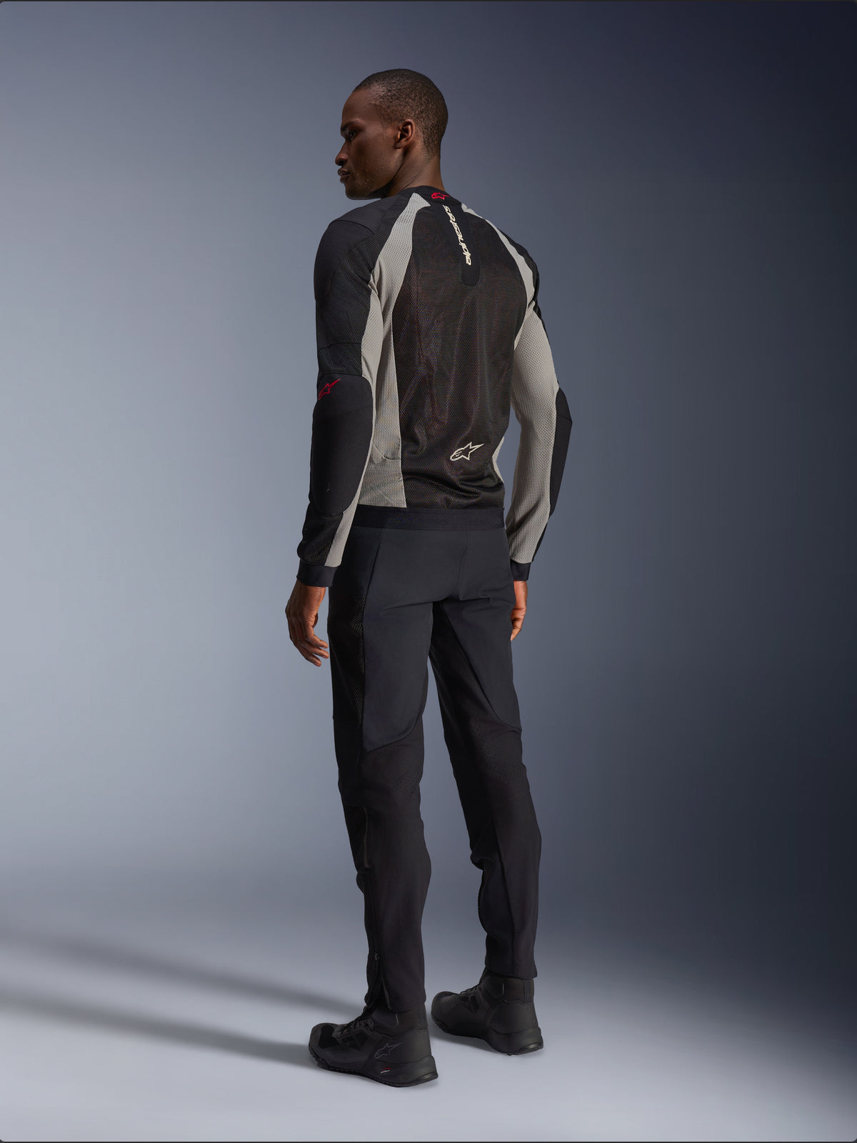 Modelo luciendo la chaqueta Alpinestars Troop-Air vista desde atrás, en color negro y gris ceniza con detalles en rojo brillante, construcción de malla técnica para ventilación, combinada con pantalones de moto negros y botas de conducción, estilo urbano contemporáneo