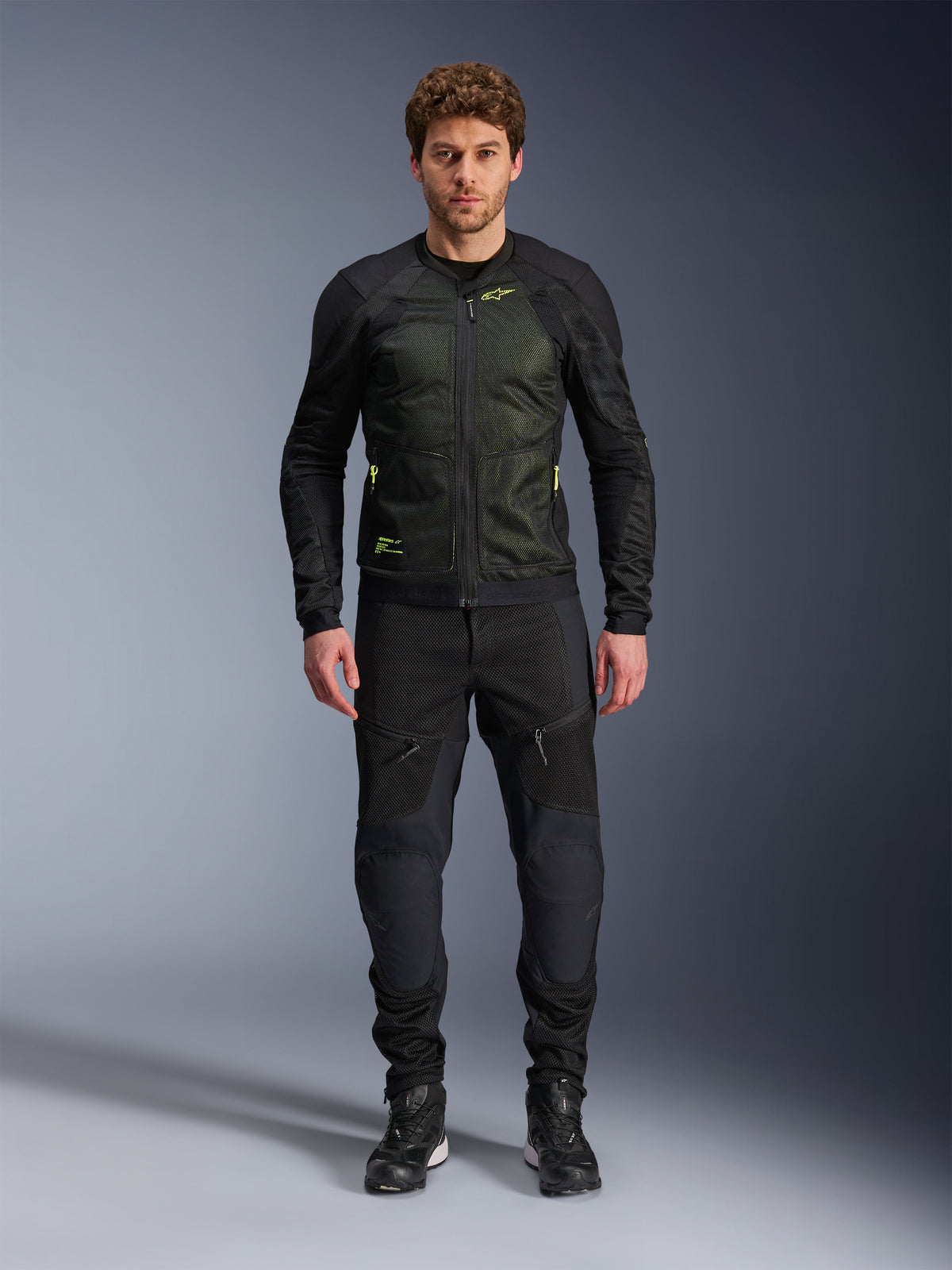 Modelo masculino vistiendo la chaqueta Alpinestars Troop-Air en Negro Amarillo Flúor, con paneles de malla técnica y detalles en amarillo flúor, combinada con pantalones y calzado de motociclismo negros, equipamiento contemporáneo para climas cálidos