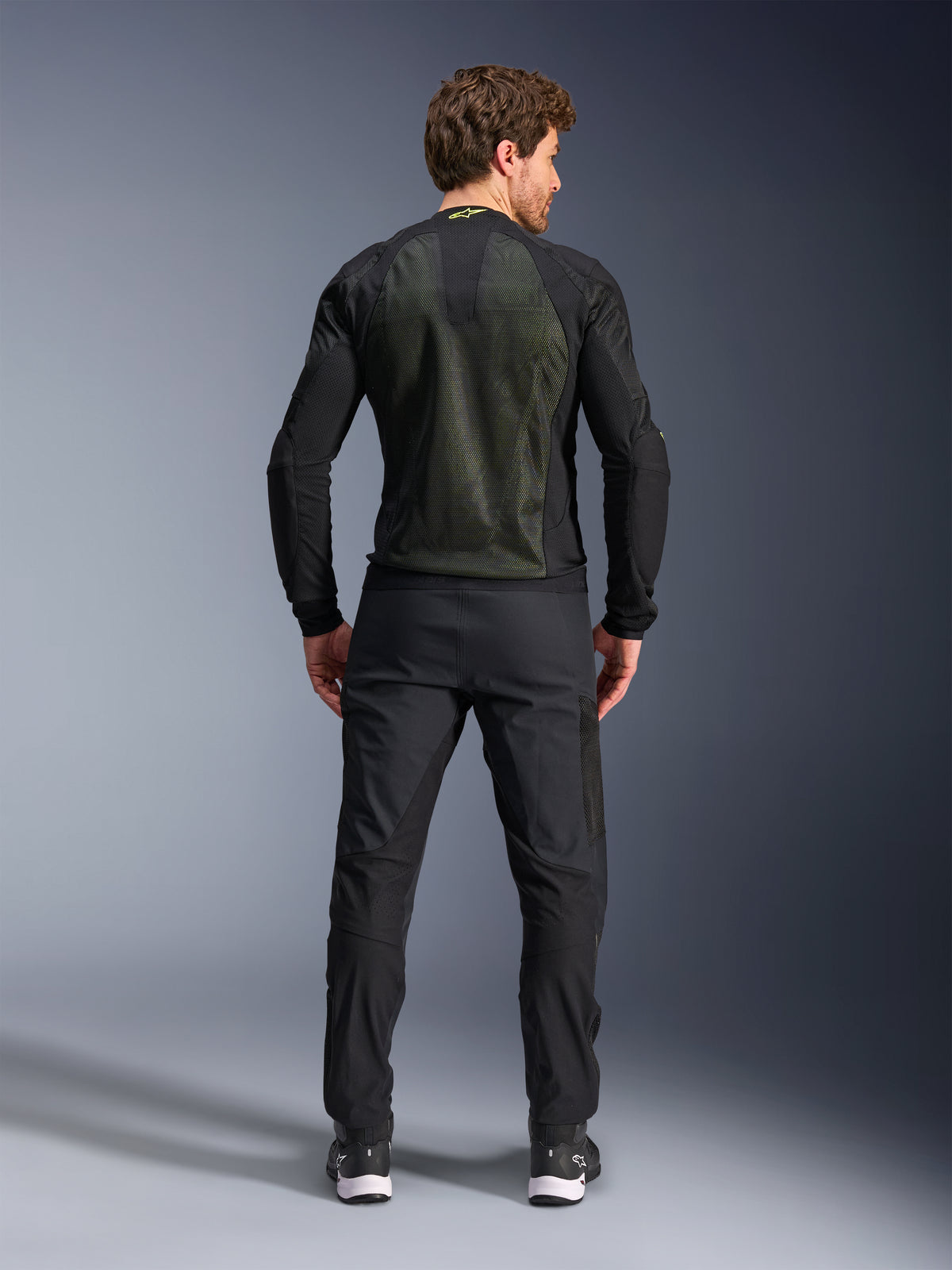 Chaqueta Alpinestars Troop-Air, Negro Amarillo Flúor, vista trasera en un modelo, con un gran panel posterior de malla técnica 2D para ventilación, chaqueta técnica de motociclismo usada con pantalones y botas de conducción negros, estilo de moto urbano