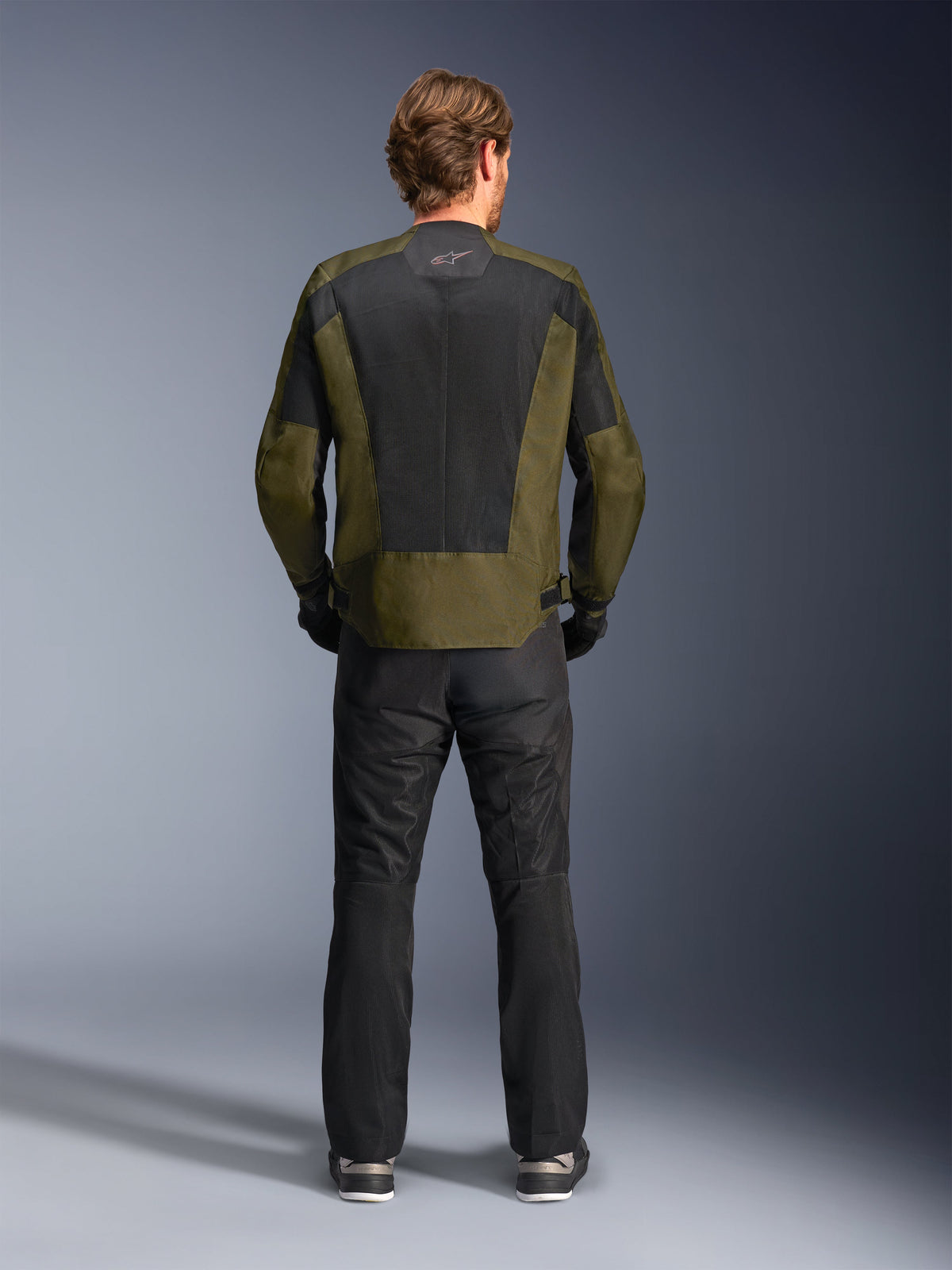 Vista posterior de un motociclista con la chaqueta Alpinestars C-1 Air en color caqui y negro, con un gran panel de malla técnica en la espalda para ventilación, combinada con pantalones de moto negros, equipamiento de conducción urbana contemporánea.