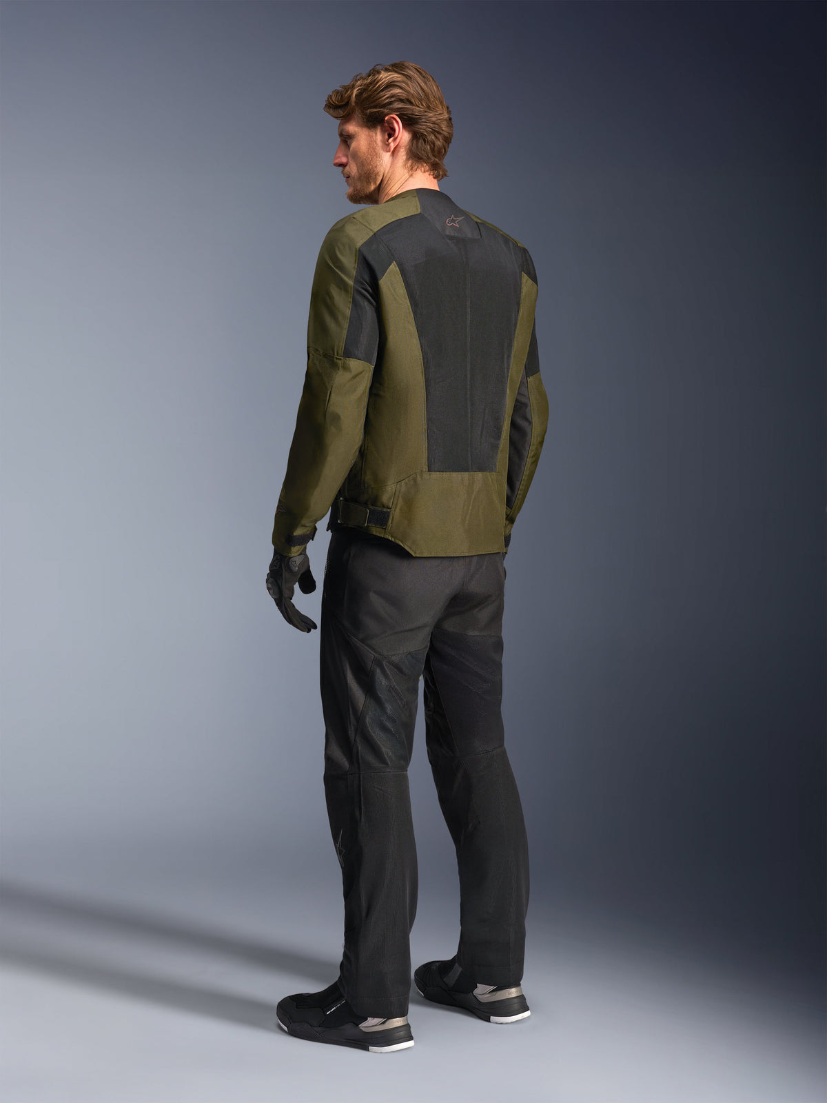 Modelo masculino con la chaqueta Alpinestars C-1 Air en color kaki, chaqueta de moto urbana, vista trasera destacando los grandes paneles de malla negra en la espalda, combinada con pantalones técnicos de conducción y guantes negros, equipamiento de moto contemporáneo para clima cálido