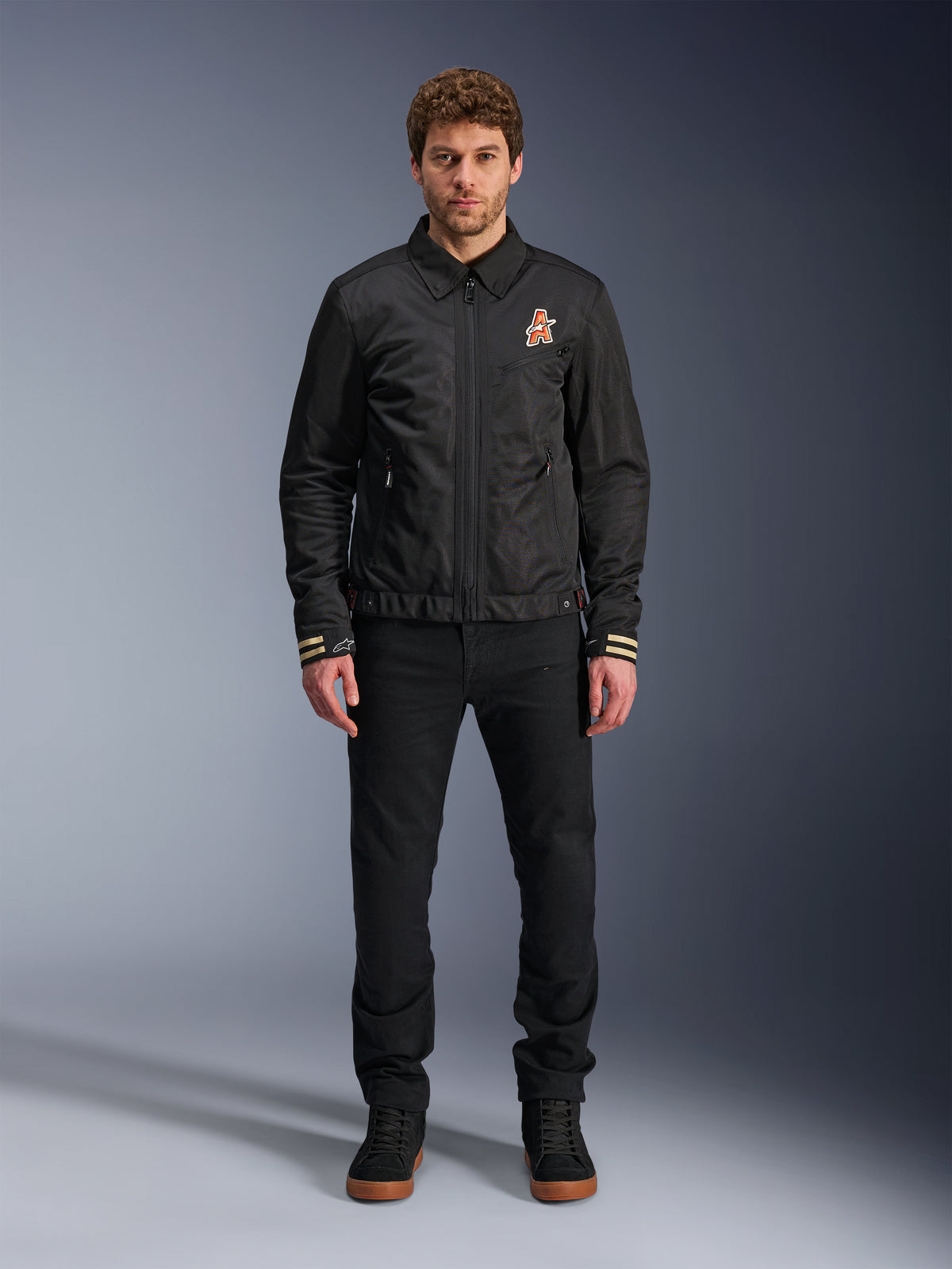 Modelo masculino vistiendo la chaqueta Alpinestars Ignite Air en negro, con cuello de camisa clásico, cremallera frontal y un logotipo naranja en el pecho, combinada con pantalones negros y zapatillas altas negras para un estilo de motociclista urbano contemporáneo.