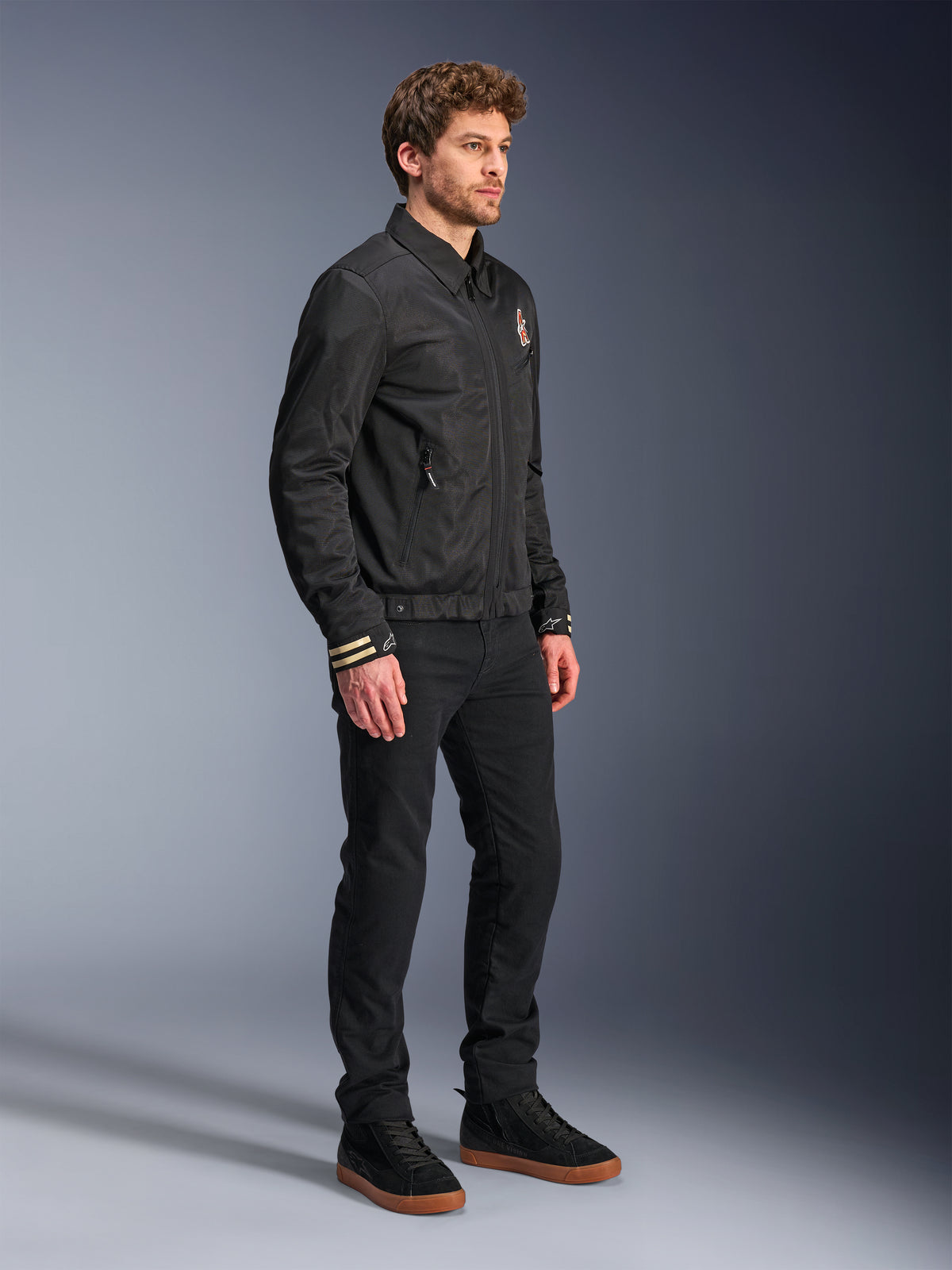 Chaqueta Alpinestars Ignite Air, chaqueta de moto urbana, negra, usada por un modelo con pantalones negros y zapatillas negras, con un cuello de camisa tradicional, gráficos inspirados en clubes en el pecho y los puños, diseño contemporáneo elegante con acabado de malla y satén