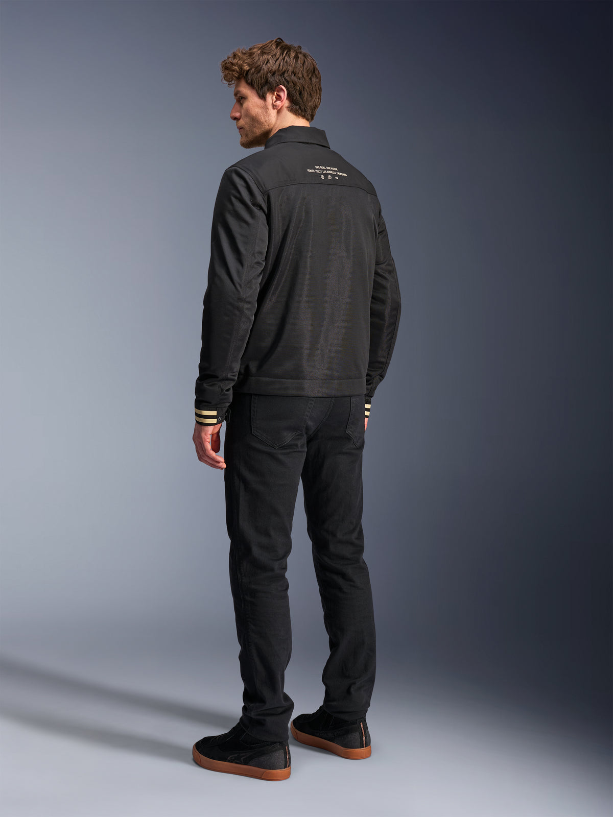 Chaqueta Alpinestars Ignite Air, chaqueta de moto negra, vista posterior con texto gráfico blanco en la parte superior de la espalda y puños con rayas color canela, silueta estilo Harrington con cuerpo de malla, combinada con pantalones negros y zapatillas negras