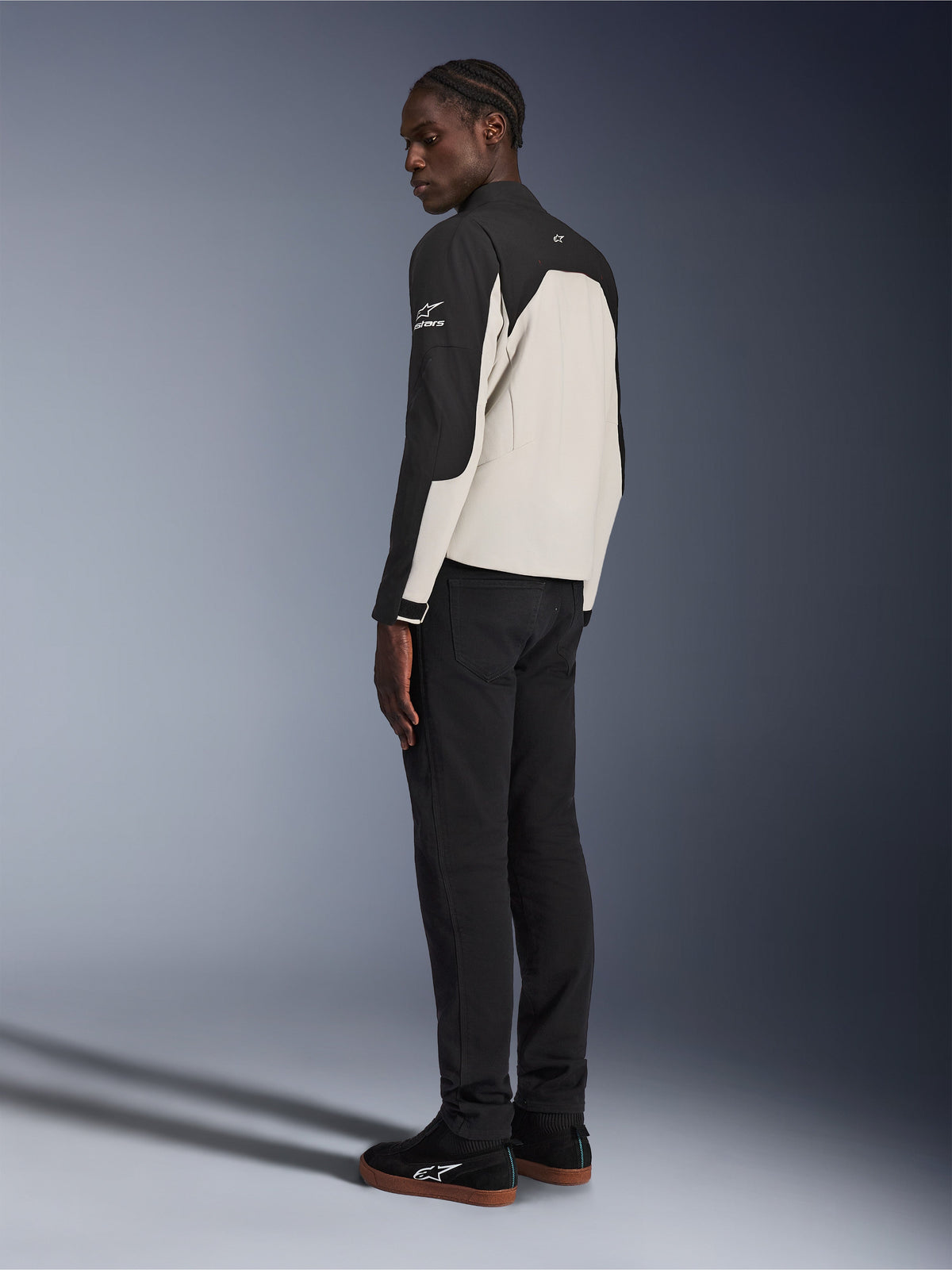 Chaqueta Alpinestars Provoke, chaqueta de moto urbana, negro y gris metalizado, vista trasera de tres cuartos en un modelo, construcción textil bitono con el logotipo de Alpinestars en el hombro, combinada con pantalones negros y zapatillas negras, protección urbana contemporánea