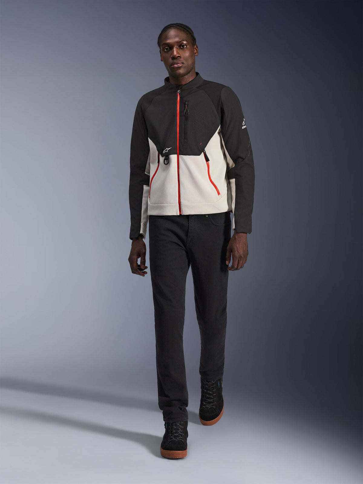 Modelo masculino vistiendo la chaqueta Alpinestars Provoke, chaqueta de moto urbana en negro y gris metalizado con detalles de cremalleras en rojo, combinada con pantalones negros y zapatillas altas negras, equipo de protección moderno de estilo urbano para conducción en ciudad