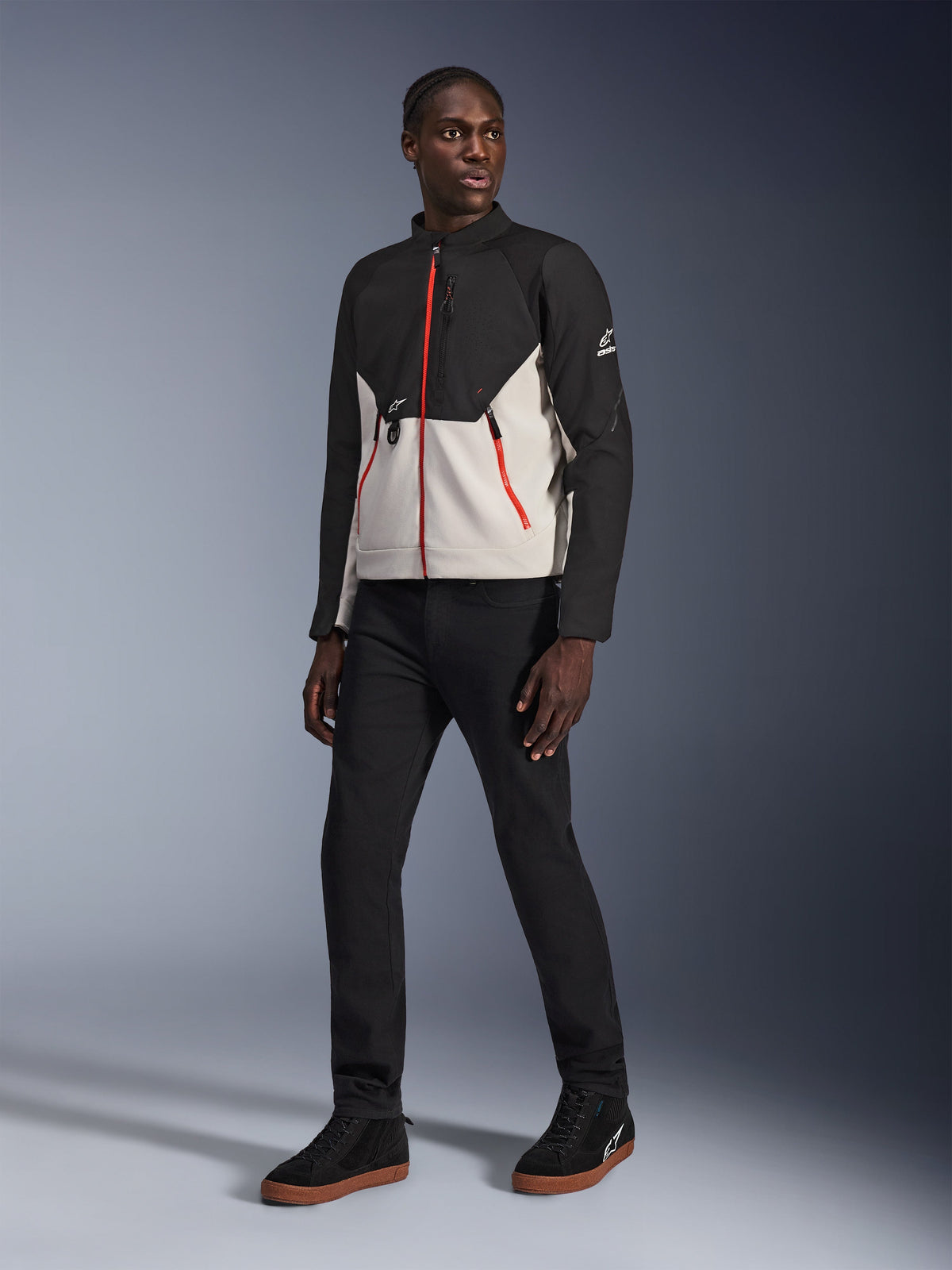 Modelo vistiendo la chaqueta Alpinestars Provoke en color Black Metal Gray, con distintivas cremalleras rojas frontales y en los bolsillos, combinada con pantalones negros y zapatillas negras de caña alta con suelas de goma color caramelo, equipamiento de motociclismo urbano contemporáneo para un look listo para la calle.