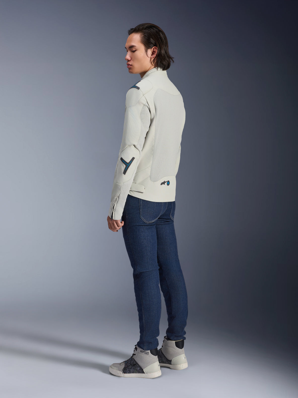 Chaqueta Alpinestars Circal-1 Air, chaqueta de moto urbana, Gris Metalizado con acentos en Azul Methyl, mostrada por un modelo en una vista de tres cuartos trasera con vaqueros azul oscuro y zapatillas altas grises, con paneles de malla y un diseño minimalista