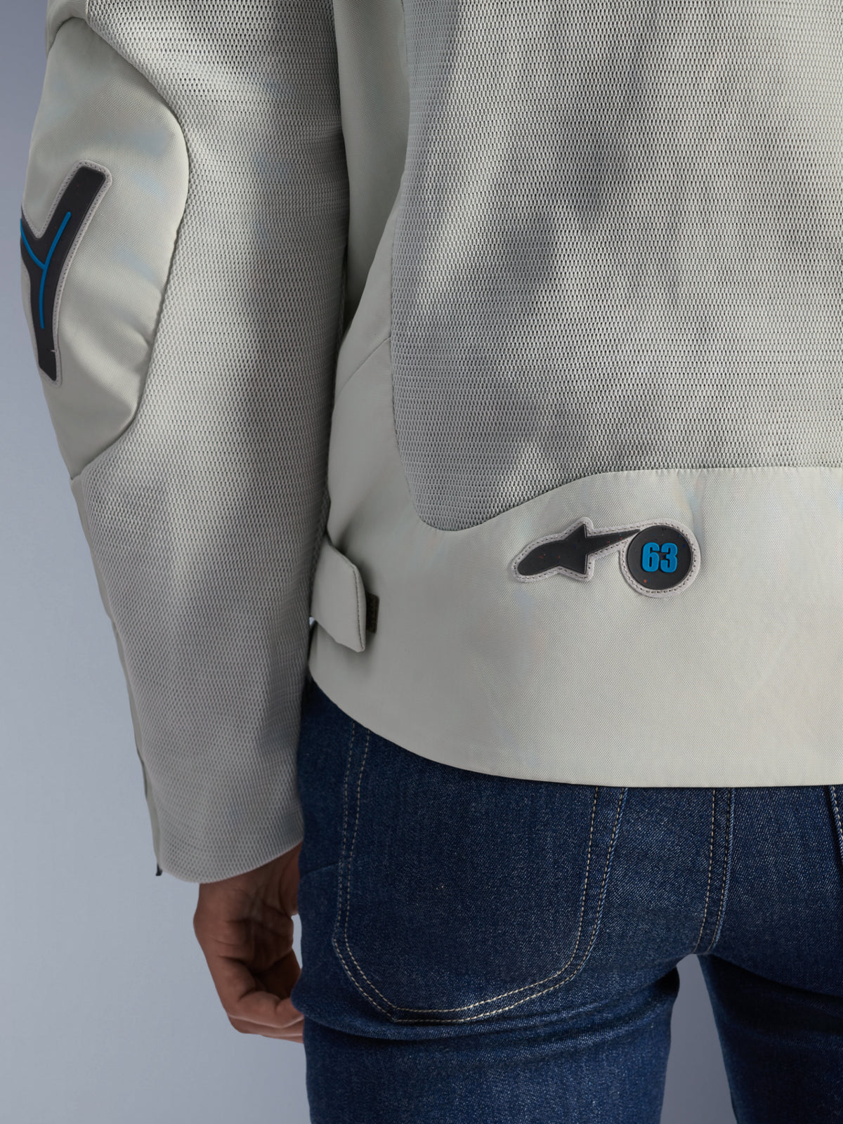 Chaqueta Alpinestars Circal-1 Air en Metal Gray con detalles en Methyl Blue, vista posterior detallada que resalta los paneles de flujo de aire de malla reciclada y un parche circular con el logotipo '63', combinada con jeans azules.