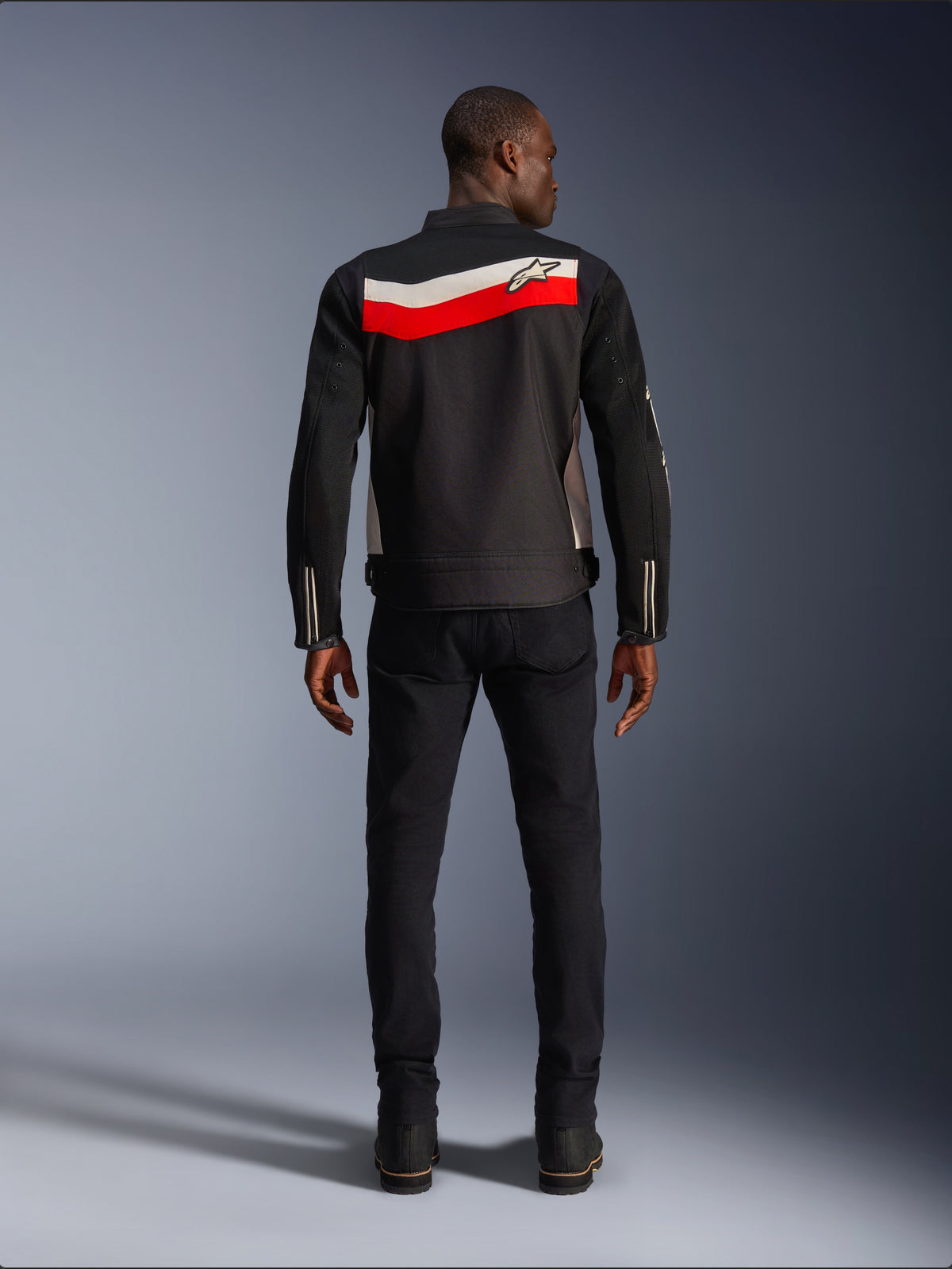 Chaqueta Alpinestars T-Dyno Air, chaqueta de moto textil para hombre, vista trasera, negra con franjas horizontales en rojo brillante y crudo en la espalda, logo de Alpinestars en el canesú, combinada con pantalones y botas negras, construcción de malla 3D transpirable