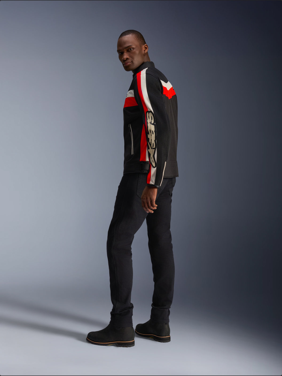 Un hombre vistiendo la chaqueta Alpinestars T-Dyno Air en negro, rojo brillante y crudo, chaqueta de moto textil de inspiración vintage con paneles de malla, marca llamativa en la manga, combinada con pantalones negros y botas para un look de motociclista moderno