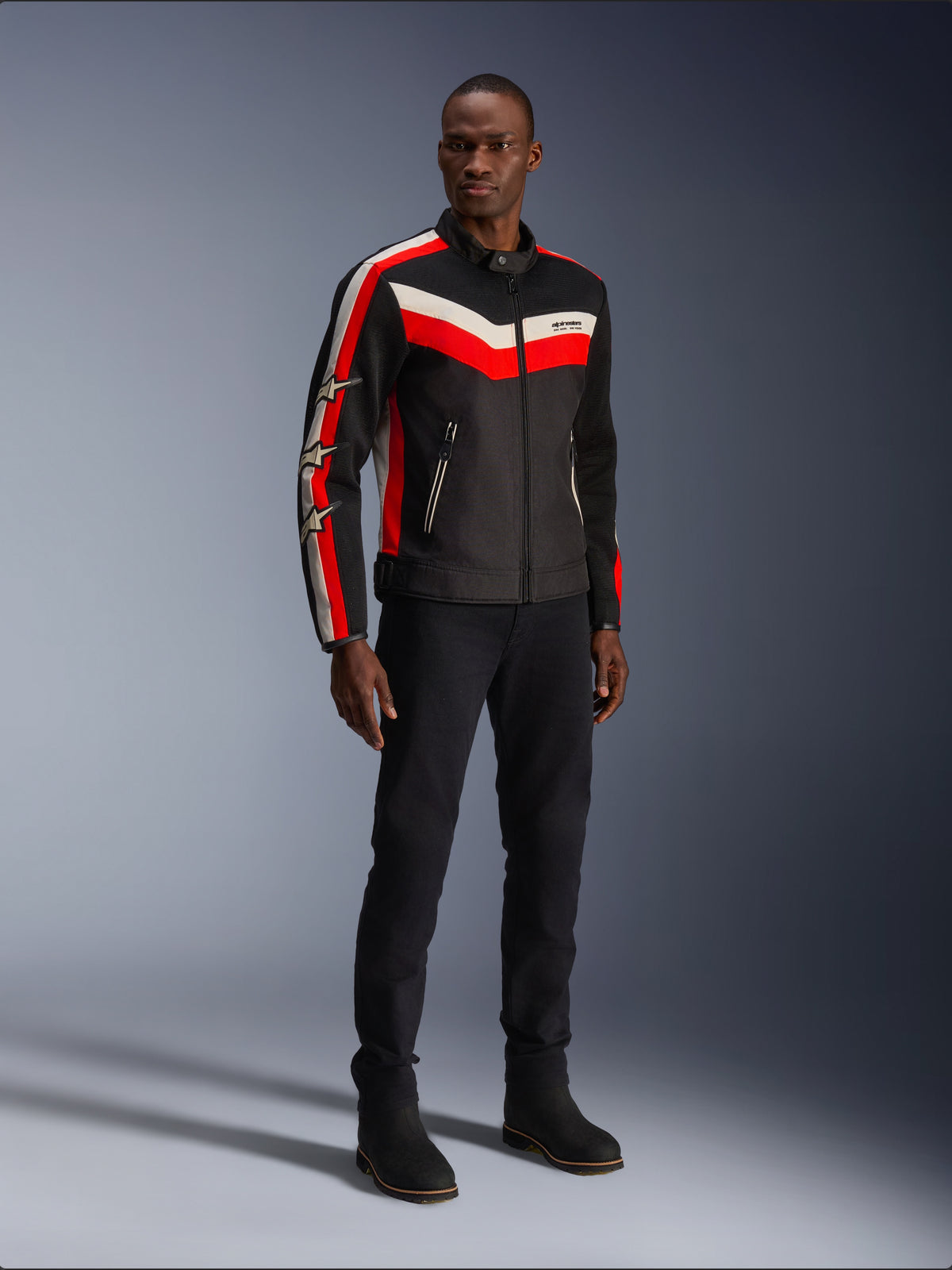Chaqueta Alpinestars T-Dyno Air, Negro Rojo Brillante Crudo, chaqueta textil de moto para hombre con paneles de malla 3D, con franjas en forma de chevron rojas y blancas en el pecho y logotipos de estrellas en las mangas, usada por un modelo con pantalones y botas negros