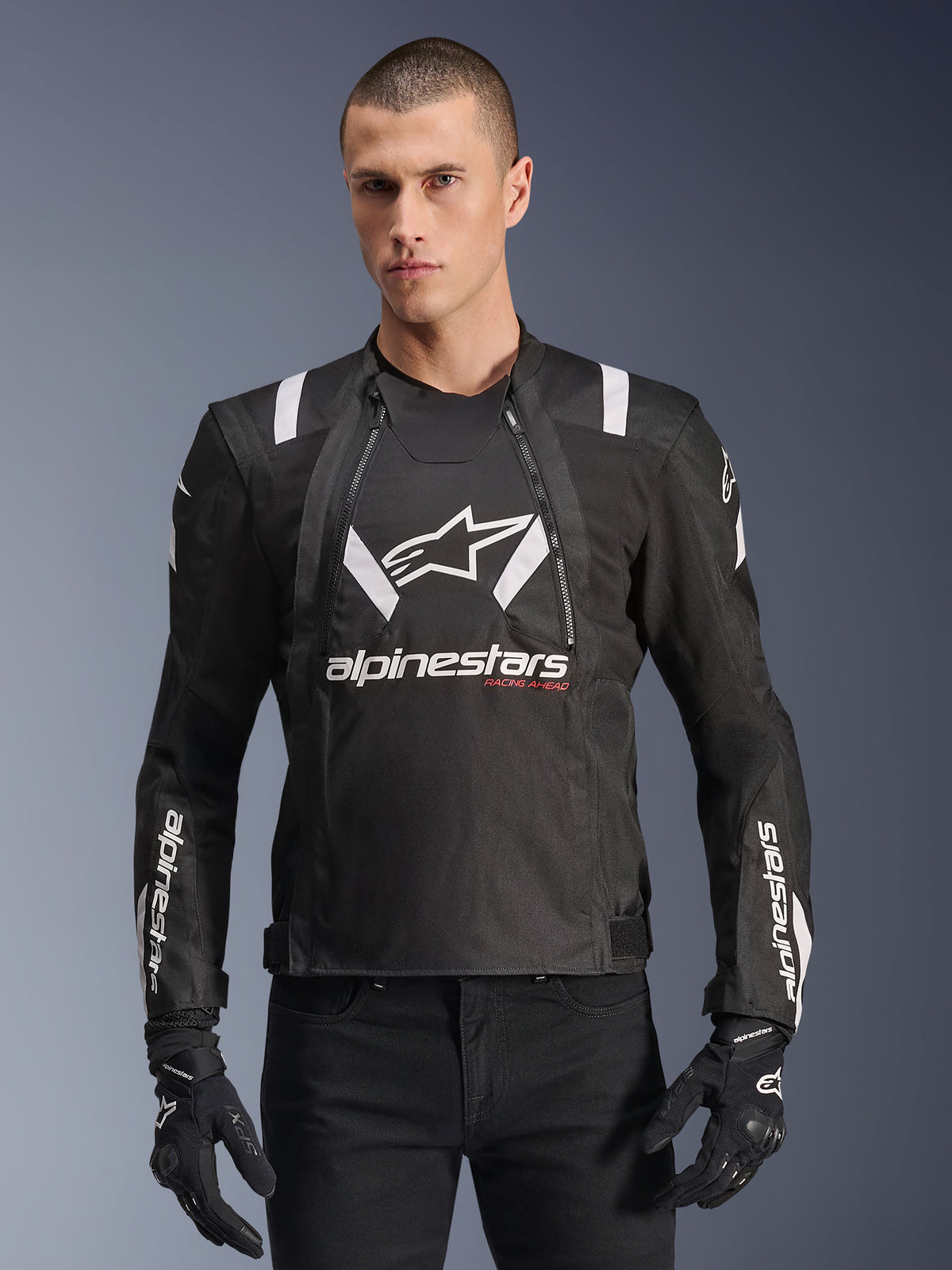 Alpinestars T-Stunt Air Jacket, Black and White Motorcycle Jacket, worn by a man with black gloves and pants, featuring bold white branding on the chest and sleeves with red 'Racing Ahead' accents, sleek sporty design with white shoulder stripes and breathable mesh panels.