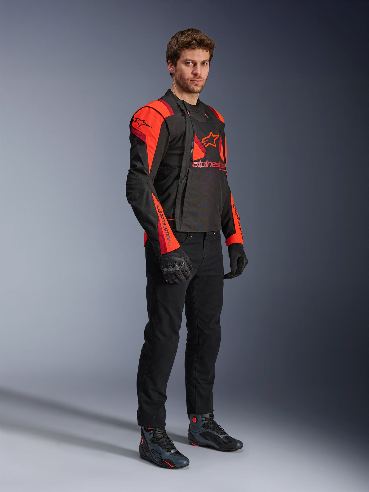 Chaqueta Alpinestars T-Stunt Air, negro, rojo flúor y rojo oscuro, chaqueta modular de motociclismo usada por un modelo con los paneles frontales abiertos para mostrar el interior de malla, combinada con pantalones, guantes y calzado de moto negros.
