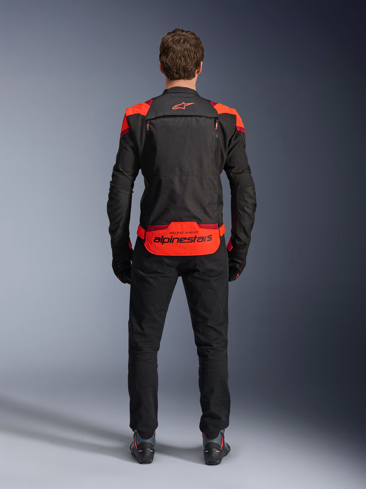 Chaqueta Alpinestars T-Stunt Air, negra con detalles en rojo flúor y rojo oscuro, vista posterior en modelo, combinada con pantalones y botas de moto negros, con un llamativo logotipo de Alpinestars en el panel inferior de la espalda, diseño modular con construcción de malla para ventilación.