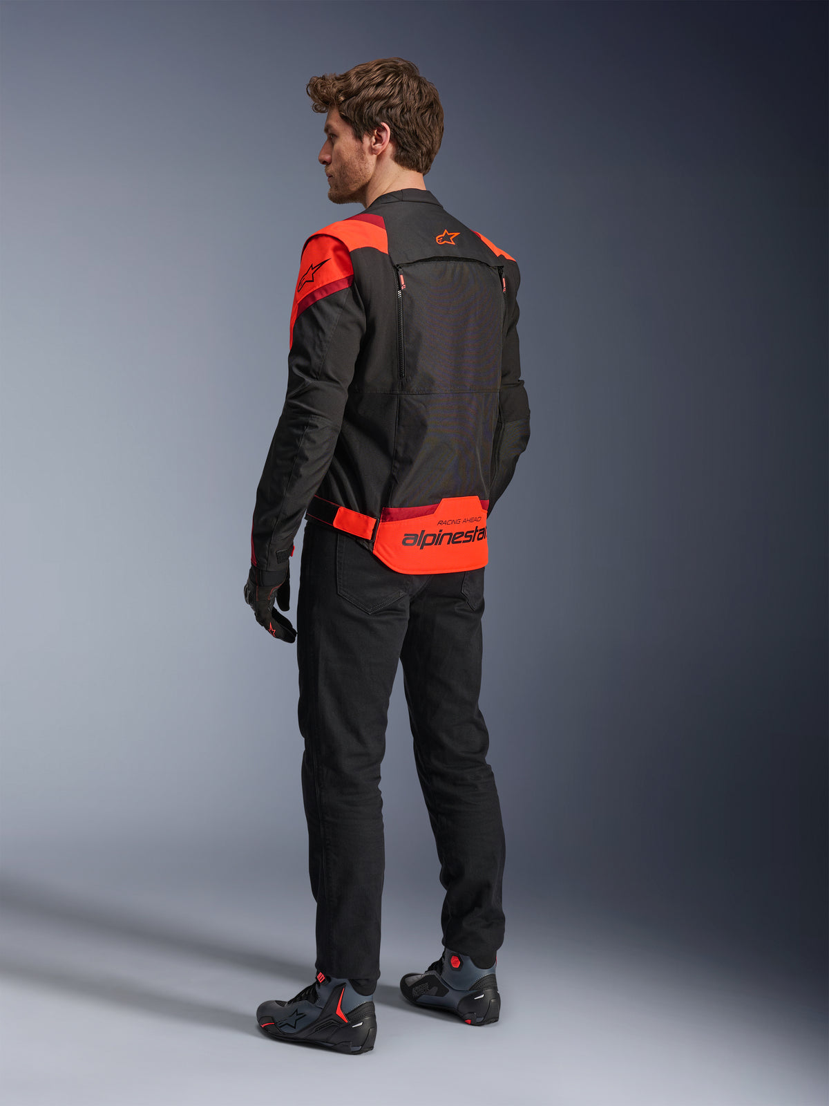 Hombre con la chaqueta Alpinestars T-Stunt Air vista desde atrás, negro, rojo fluo y rojo oscuro, chaqueta de moto textil con paneles de malla y diseño de corte deportivo, con un llamativo logotipo de Alpinestars en la parte baja de la espalda, combinada con pantalones negros y botas de motociclista