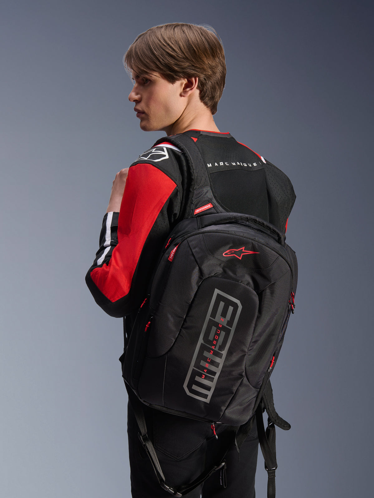 Model wearing the Alpinestars MM93 T-SPS Air V2 Jacket in Black Bright Red and White, viewed from the back while carrying a black Alpinestars backpack with Marc Márquez branding, showcasing urban motorcycle riding apparel with mesh panels and logo details