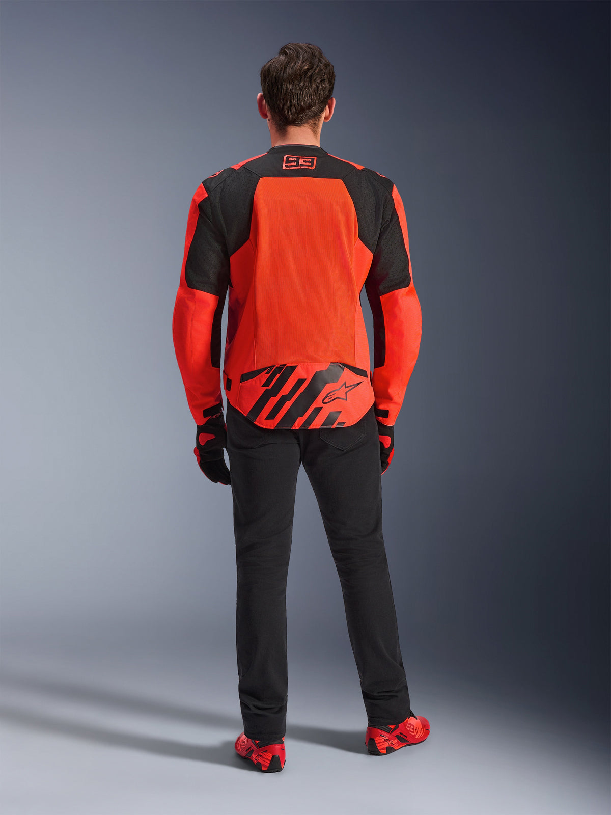Back view of the MM93 T-SPS Air V2 Jacket, Red Fluo and Black, textile motorcycle jacket with large mesh panels on the back for ventilation, worn with black pants and red footwear, featuring Marc Marquez 93 branding