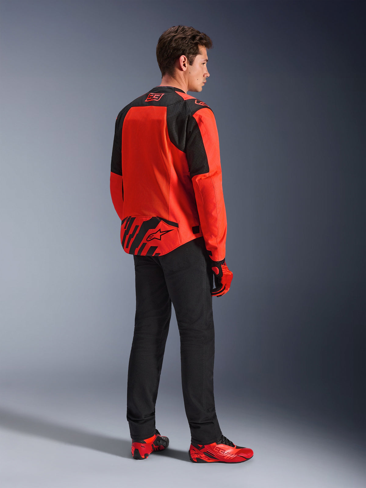 Alpinestars MM93 T-SPS Air V2 Jacket in Red Fluo and Black, back three-quarters view on a model, featuring Marc Marquez 93 branding on the collar and lower back, styled with black pants and red motorcycling shoes.