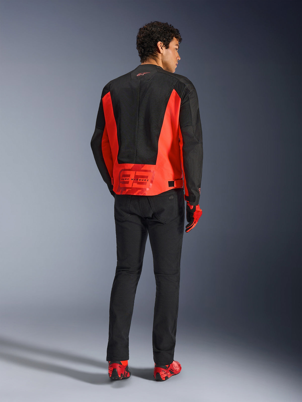 Vista trasera de un modelo masculino con la chaqueta Alpinestars MM93 C-1 Air en rojo flúor y negro, con el logotipo de Marc Márquez 93 en la parte baja de la espalda, pantalones negros y calzado de conducción rojo, estilo urbano para desplazamientos en moto