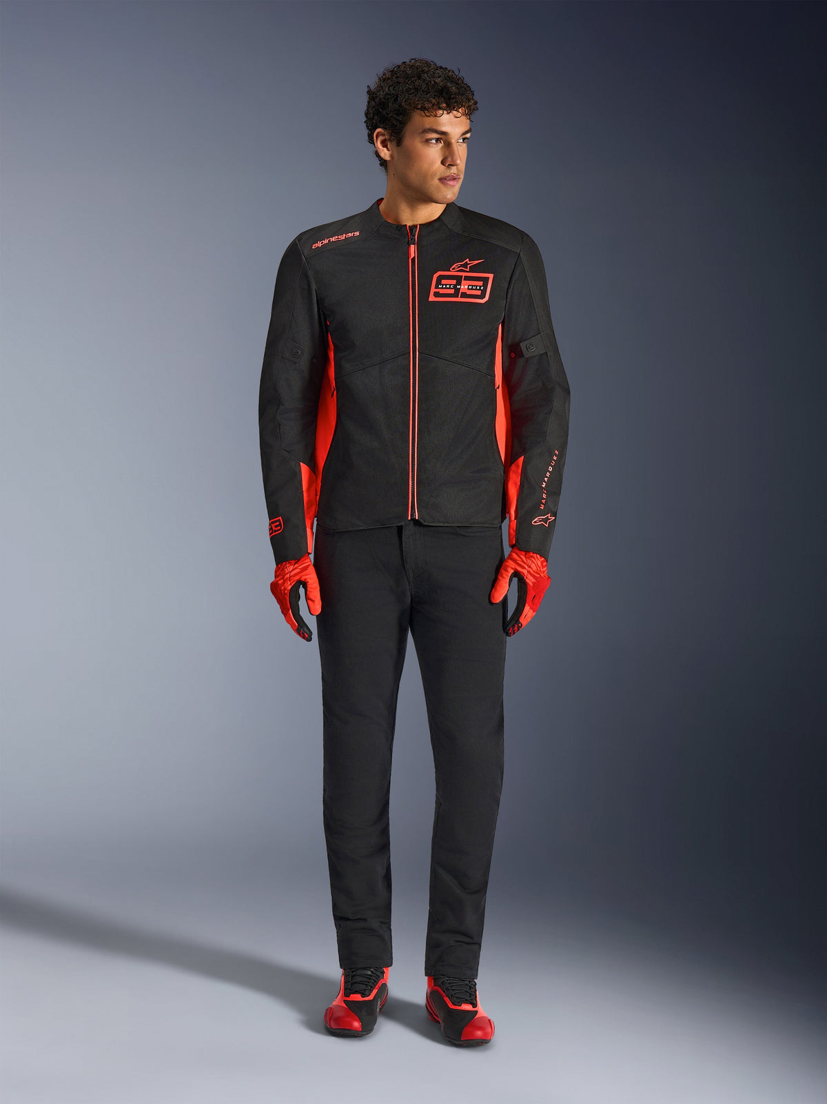 Chaqueta Alpinestars MM93 C-1 Air, chaqueta de moto, negro y rojo fluo, con la marca de Marc Márquez y el logotipo 93, usada por un modelo con guantes rojos a juego y pantalones negros, construcción ligera de textil y malla para conducción urbana