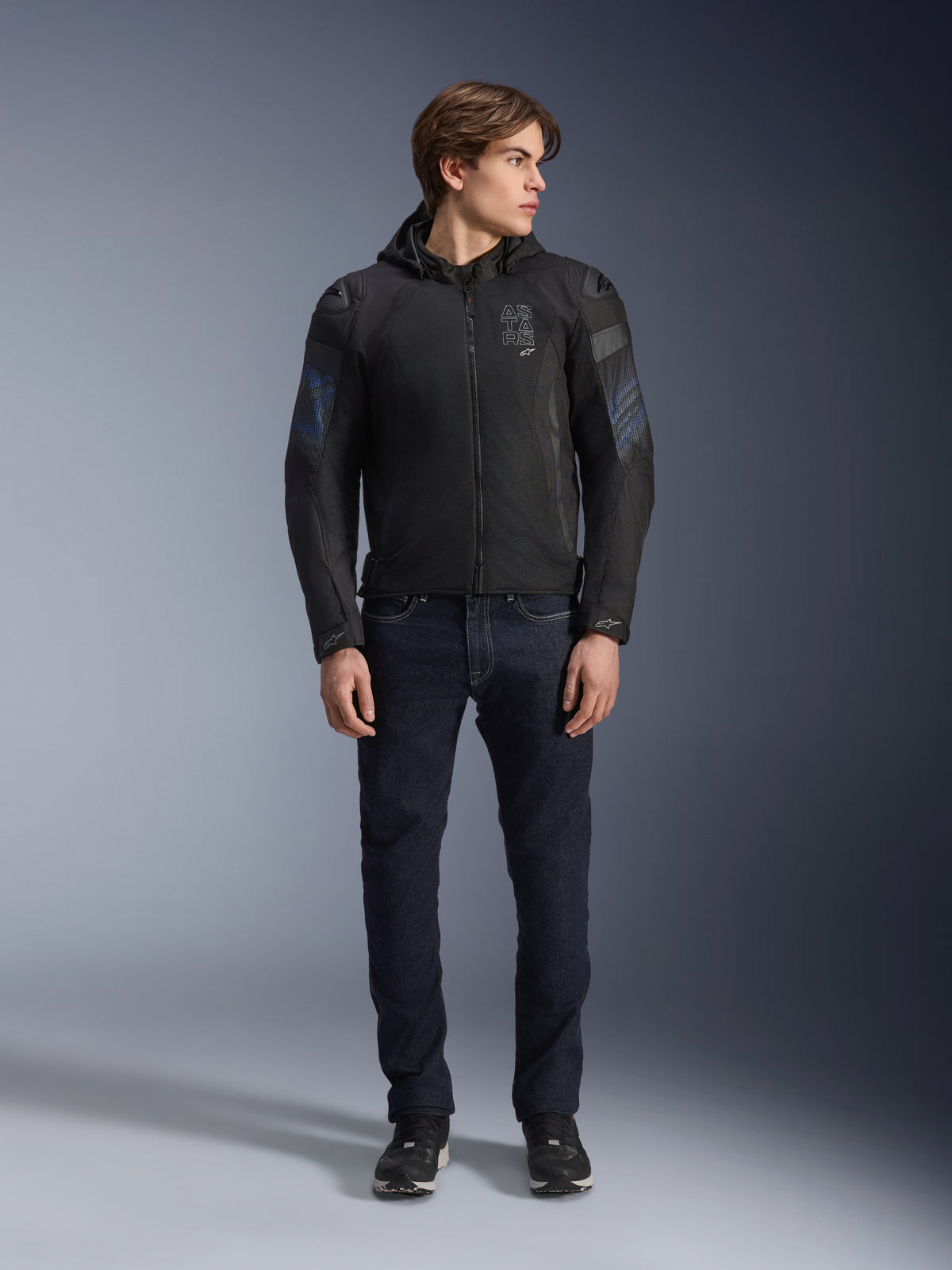 Modelo masculino con la chaqueta Alpinestars Zaca Air Venom WP en negro y azul eléctrico, chaqueta de moto textil con paneles de malla 3D y capucha desmontable, combinada con vaqueros de denim oscuro, equipamiento de conducción moderno de inspiración deportiva.