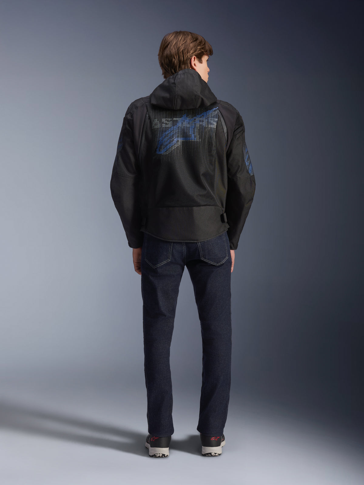 Chaqueta Alpinestars Zaca Air Venom WP, negro y azul eléctrico, vista posterior con un gran logotipo azul de Alpinestars en el panel de malla trasero, capucha desmontable, combinada con vaqueros de denim oscuro y zapatillas, equipamiento deportivo de moto para todo tipo de clima.