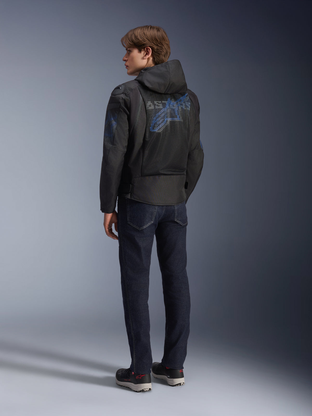 Alpinestars Zaca Air Venom WP Jacket, chaqueta de moto de tejido de malla en negro y azul eléctrico, vista trasera en un modelo, con logotipo Astar azul en la espalda y capucha desmontable, combinada con vaqueros oscuros y calzado de conducción negro.