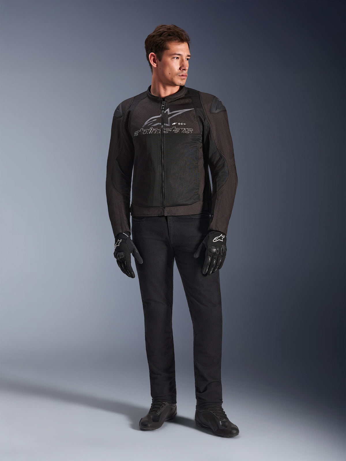 Male model wearing Alpinestars SMX Air Jacket, Black Anthracite motorcycle jacket with ventilated mesh panels and grey chest logo, paired with black pants and protective gloves, studio setting with a gradient background