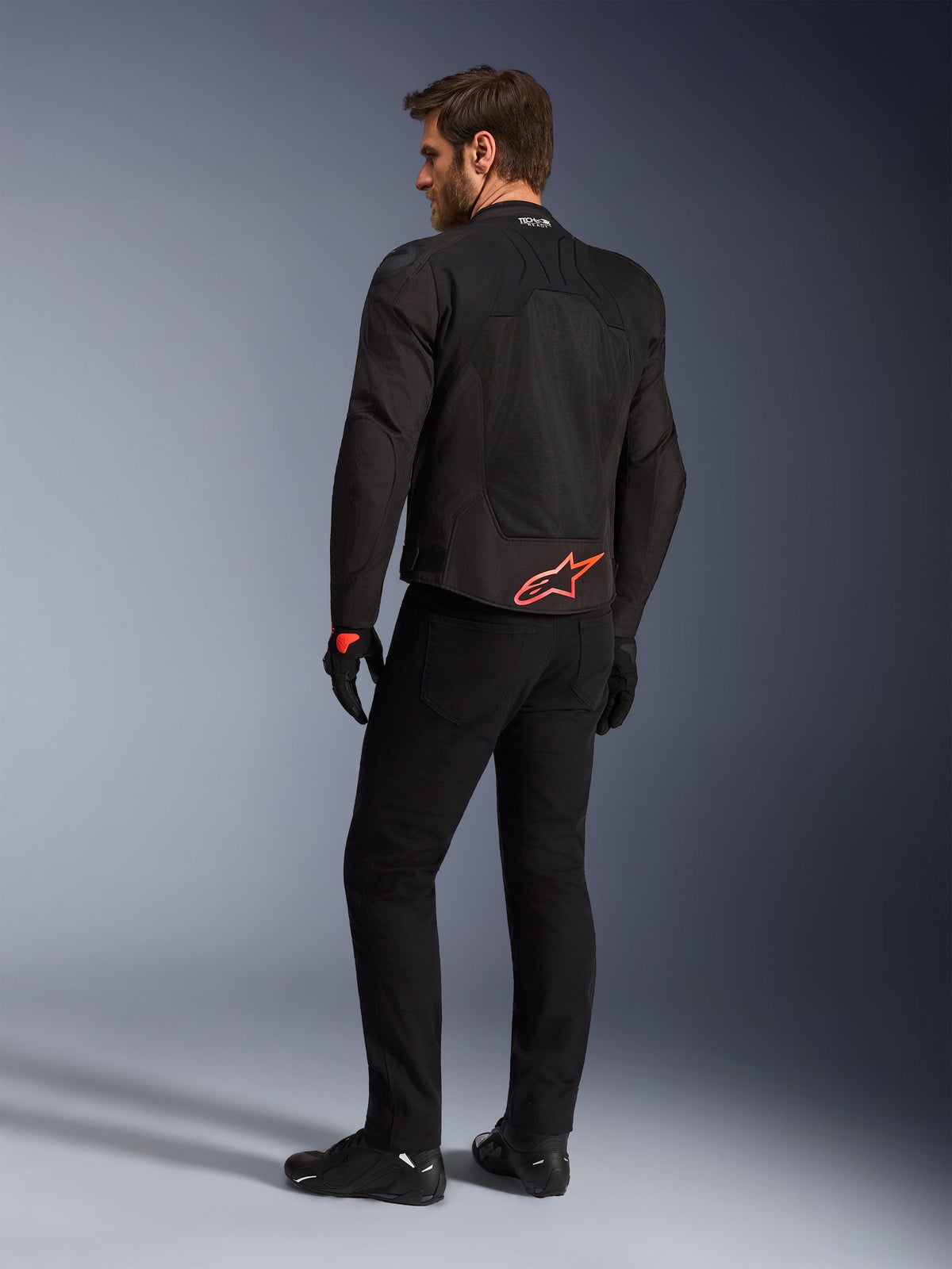 Vista trasera de un modelo luciendo la chaqueta Alpinestars SMX Air en negro, con un logotipo de Alpinestars en rojo brillante en la parte inferior de la espalda, la marca 'Tech-Air Ready' en el cuello y paneles de malla técnica para ventilación, combinada con pantalones y guantes de conducción negros.