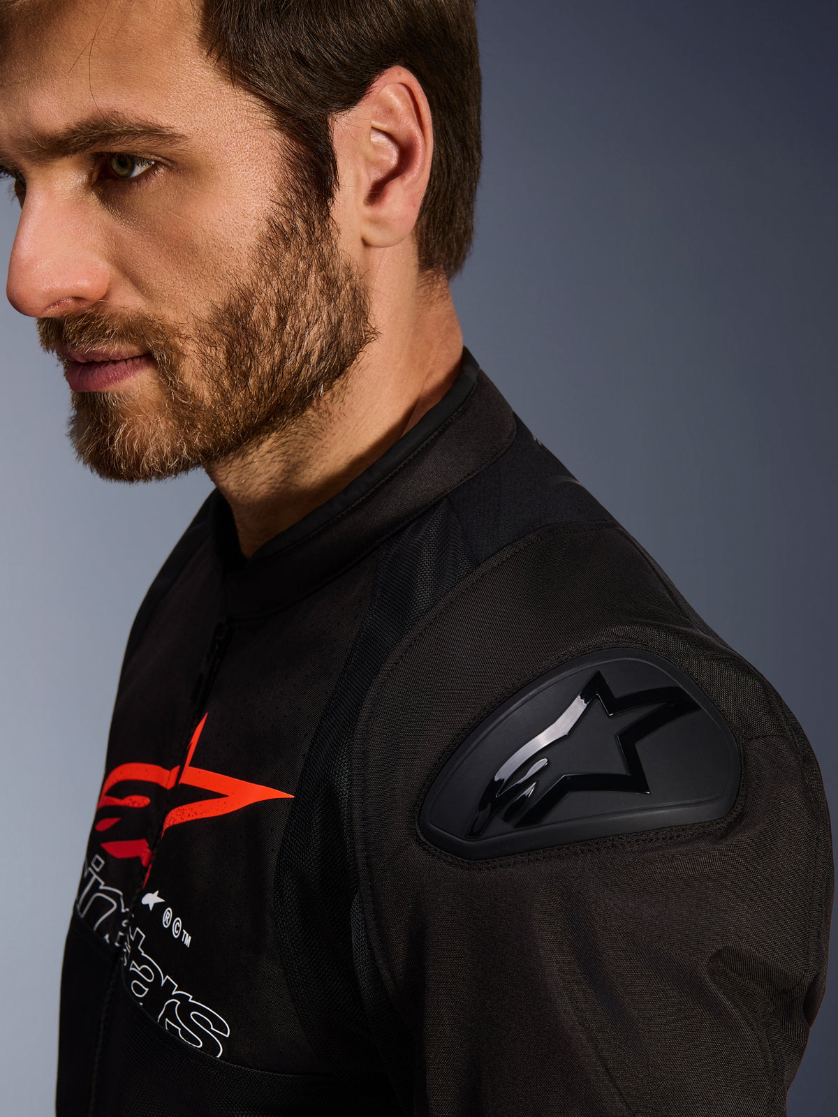 Chaqueta Alpinestars SMX Air, negra y roja, detalle en primer plano del protector de hombro DFS Lite con logo de estrella, usada por un hombre con barba, chaqueta textil de motociclismo para carreras y deportiva.