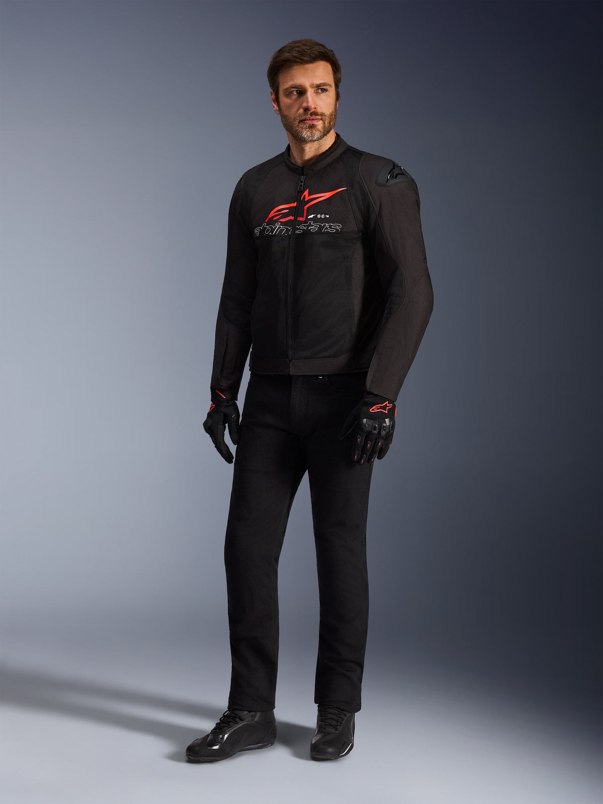 Modelo masculino vistiendo la chaqueta Alpinestars SMX Air en negro, rojo brillante y blanco, combinada con pantalones técnicos negros, guantes negros y rojos y calzado de conducción negro, con un logotipo rojo destacado en el pecho y detalles de marca en blanco, toma de estudio de cuerpo completo para equipamiento de motociclismo deportivo