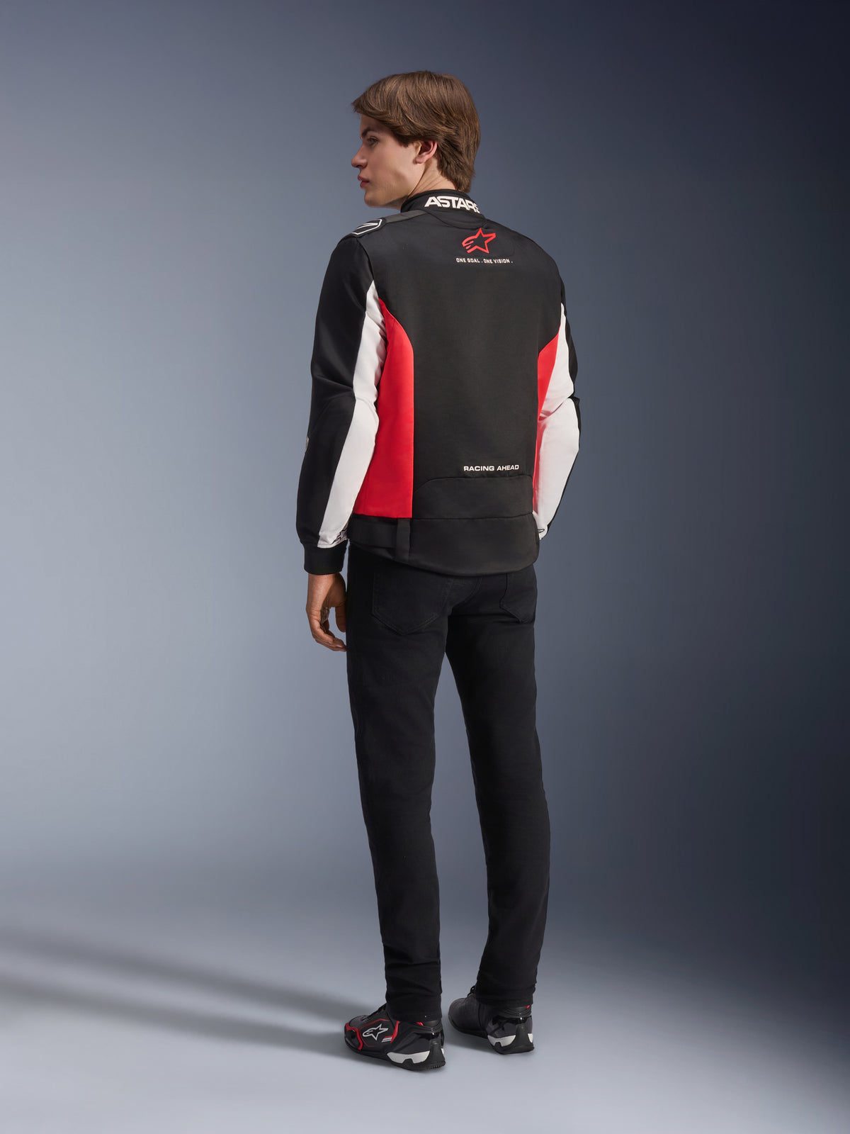 Alpinestars Monza Sport Jacket, textile motorcycle jacket, Black Bright Red and White, back view of model featuring red side panels and white sleeve accents, Alpinestars logo and 'ONE GOAL. ONE VISION.' text, paired with black pants and riding shoes for a sport-inspired look