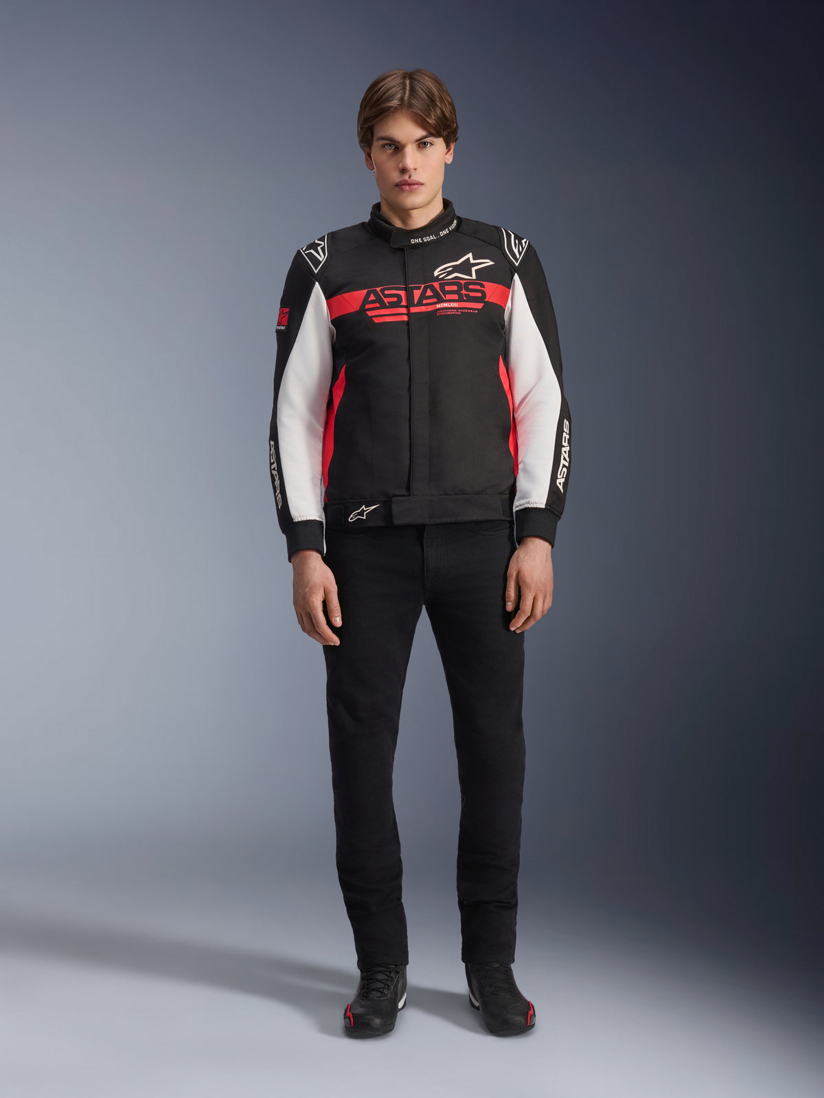 Male model wearing the Alpinestars Monza Sport Jacket in Black, Bright Red, and White, featuring a large red 'ASTARS' logo across the chest and white panels on the sleeves, paired with black pants and black riding boots.
