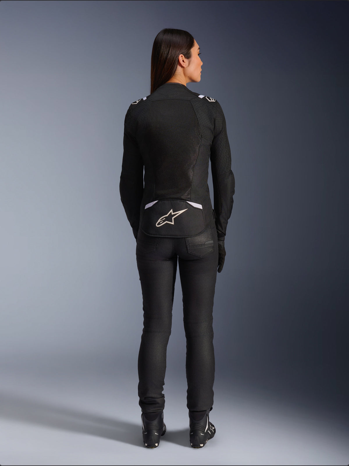 Chaqueta Alpinestars Stella T-SPS Air v2, chaqueta de moto textil para mujer, negra y blanca, vista trasera que muestra los paneles de ventilación de malla y el logotipo blanco en la parte inferior de la espalda, usada con pantalones de conducción y botas negros, equipamiento deportivo urbano.