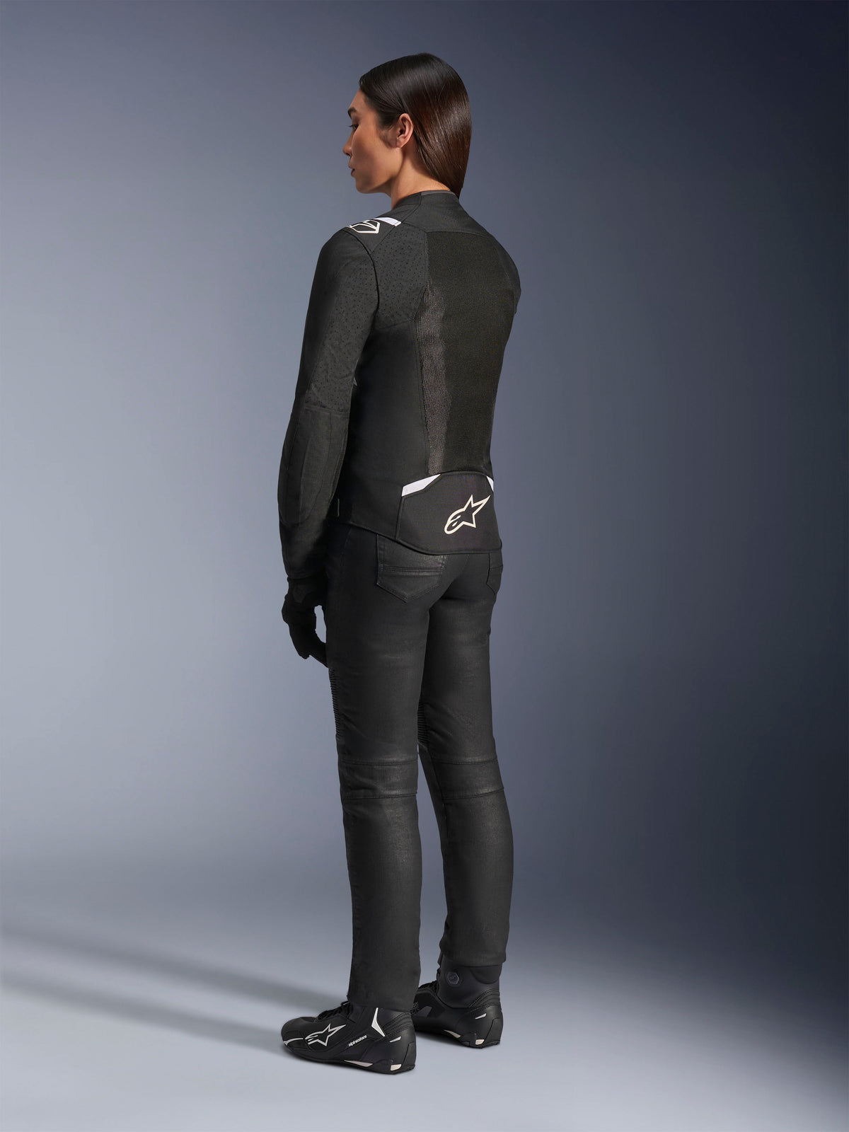 Chaqueta Alpinestars Stella T-Sps Air V2, negra y blanca, vista trasera que muestra paneles de malla y el logotipo en la parte inferior de la espalda, combinada con pantalones de conducción negros y calzado para moto, equipamiento textil deportivo de carretera para mujer