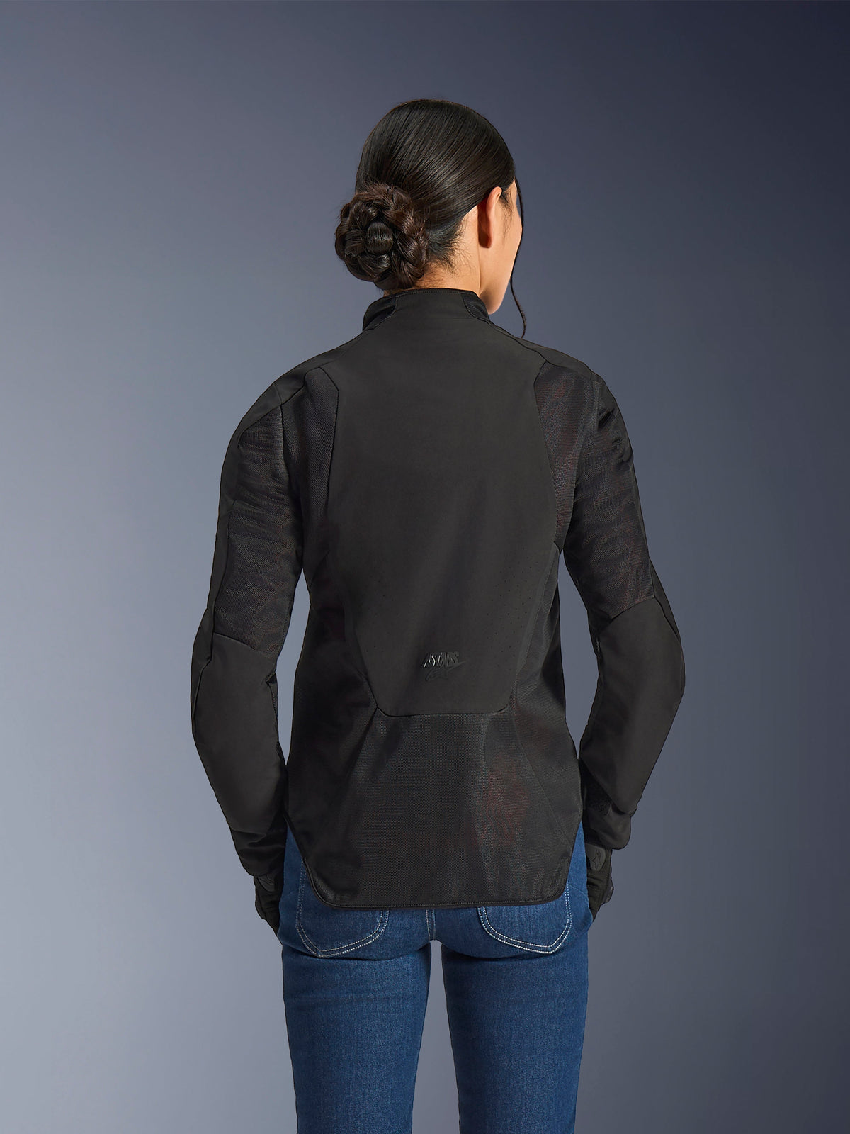 Chaqueta Stella Aeroshell Airflow de Alpinestars para mujer, Negro Antracita, vista posterior con paneles de ventilación de malla integrados en la espalda y las mangas, chasis de softshell perforado con láser y perfil trasero alargado, combinada con pantalones vaqueros azules.