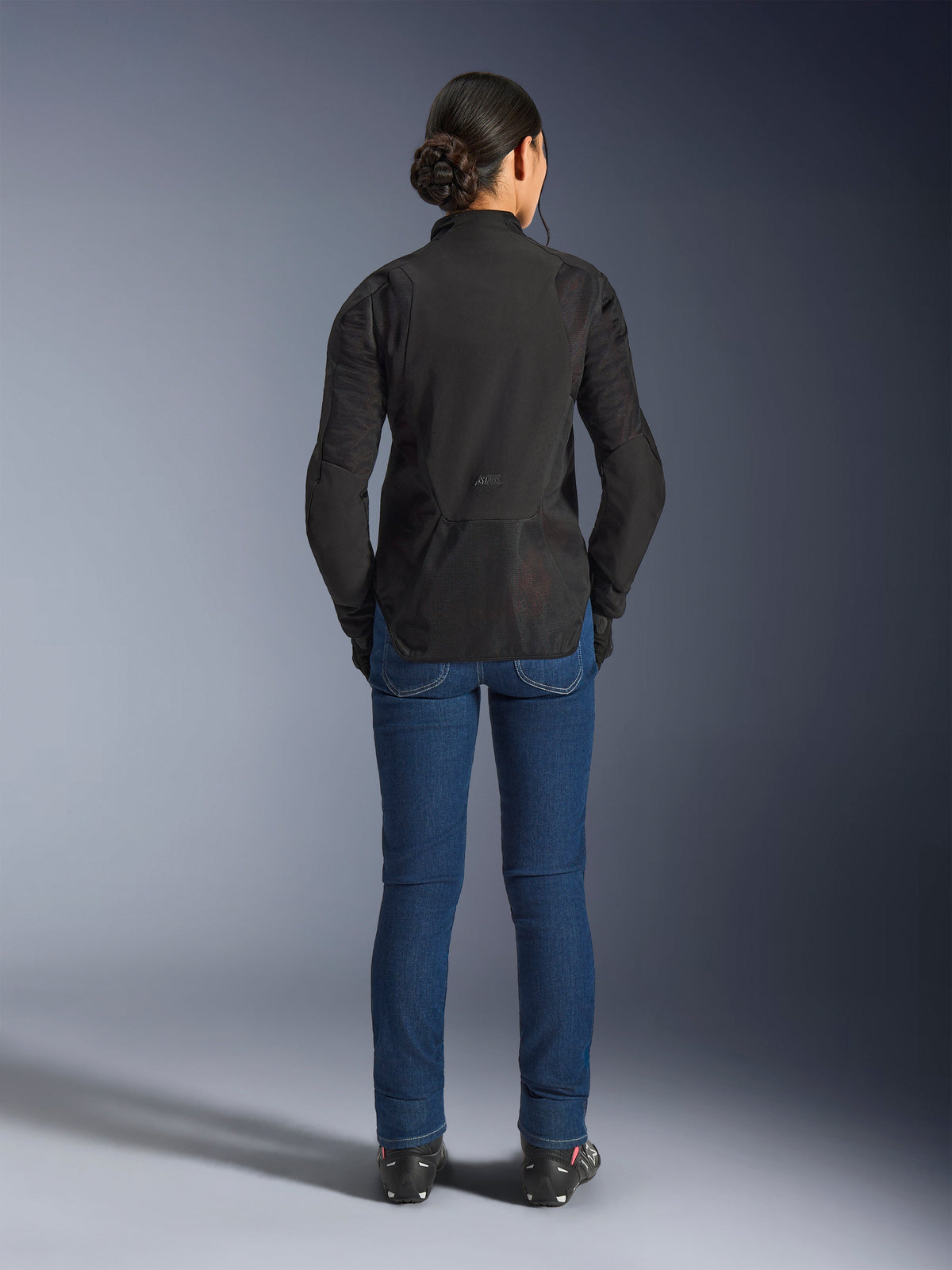 Chaqueta para mujer Stella Aeroshell Airflow de Alpinestars, color Negro Antracita, vista trasera en una modelo, con chasis de softshell ligero y paneles de malla perforados por láser, perfil trasero extendido para mayor cobertura, ajuste de conducción deportiva y combinada con vaqueros azules