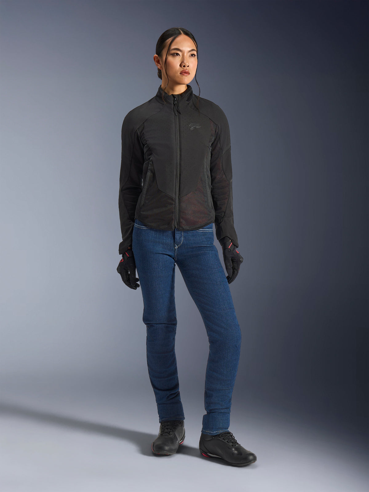 Mujer motociclista con la chaqueta Alpinestars Stella Aeroshell Airflow en color negro, con ventilación perforada con láser y paneles de malla, combinada con pantalones de moto azules y equipamiento de motociclismo negro, estética moderna de conducción deportiva y urbana