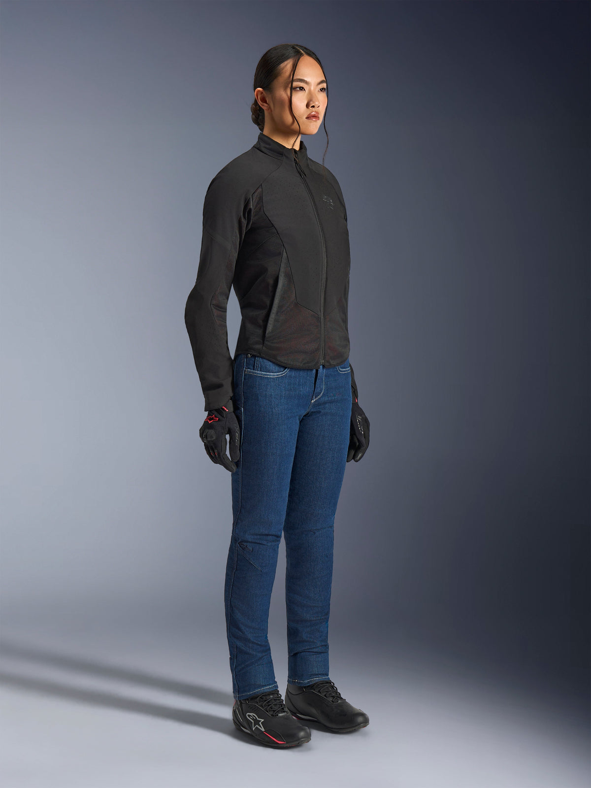 Mujer piloto vistiendo la chaqueta Alpinestars Stella Aeroshell Airflow en color Black Anthracite, chaqueta de moto softshell de corte ajustado, combinada con vaqueros de moto azules y guantes negros, vista de cuerpo completo en estudio