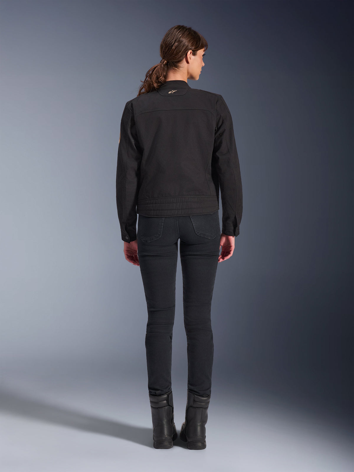 Chaqueta Alpinestars Stella Grace, chaqueta de moto urbana para mujer, negra, vista trasera que muestra el diseño inspirado en la ropa de trabajo, combinada con pantalones negros de corte ajustado y botas negras, foto de estudio